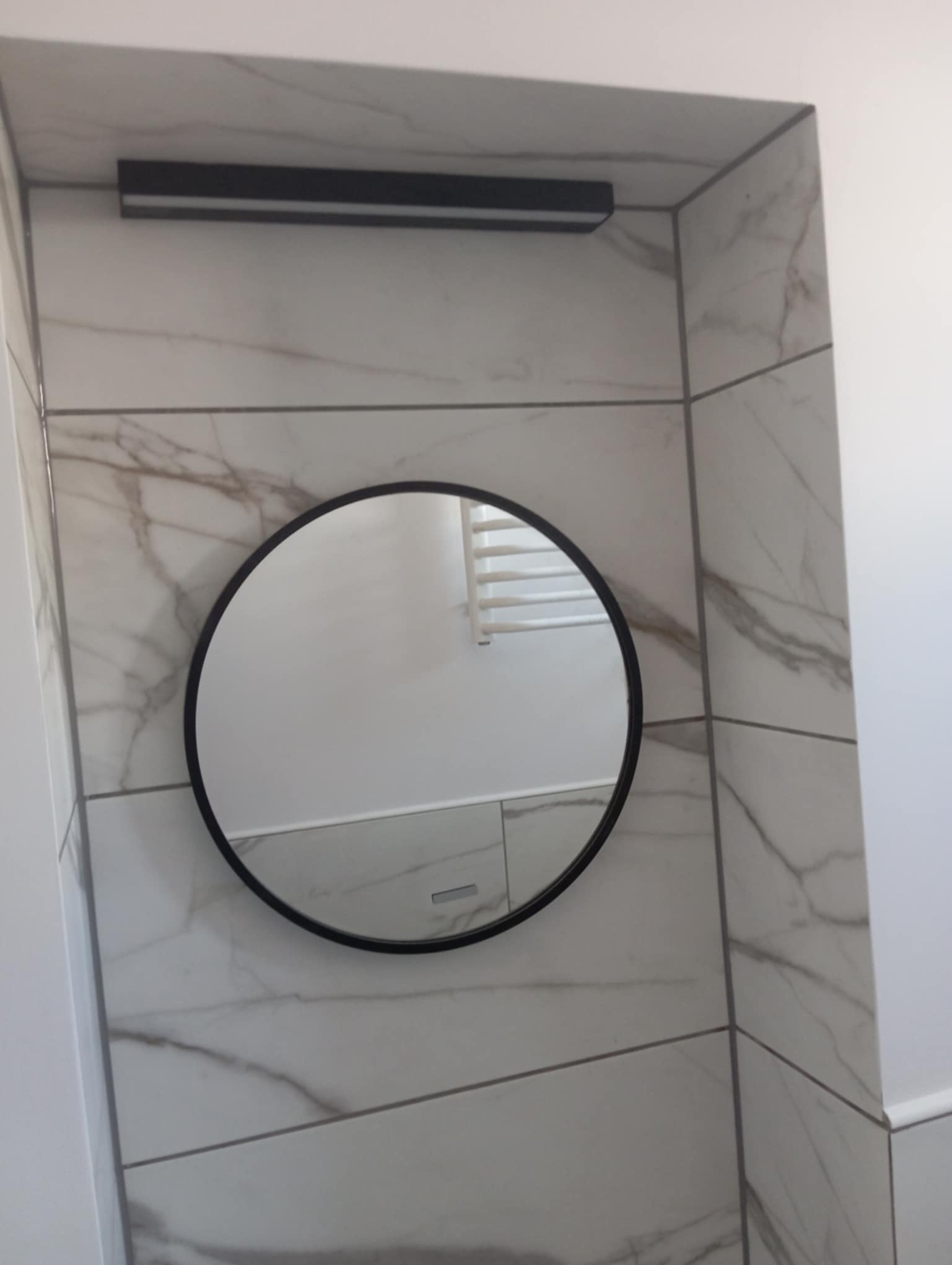 Wykończona ściana w łazience z marmurowymi płytkami, okrągłym lustrem w czarnej ramie i podłużnym oświetleniem nad nim. Widoczne fugi i odbicie grzejnika.