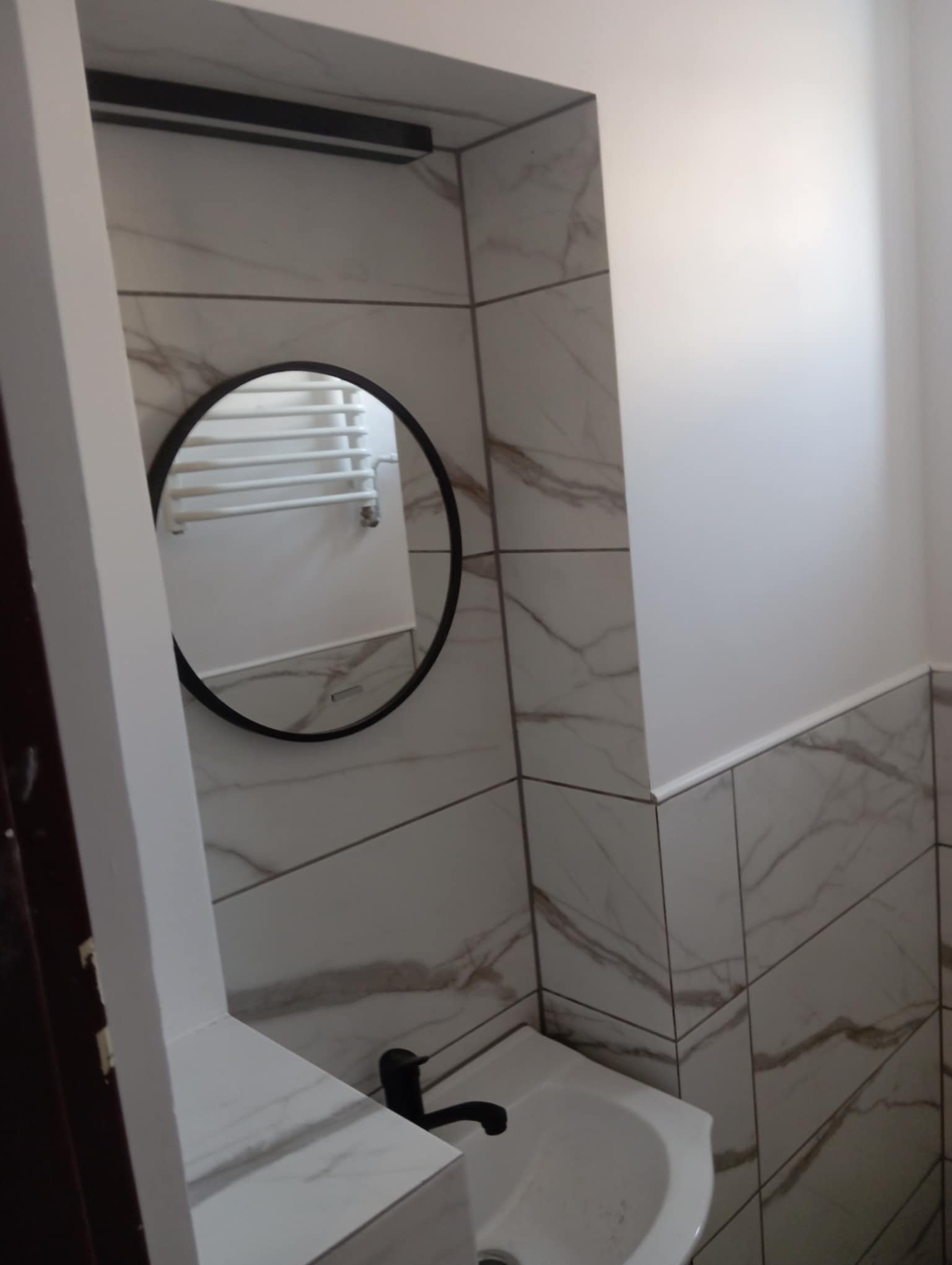 Wykończona toaleta: marmurowe płytki, okrągłe lustro z czarną ramą, biała umywalka z czarną baterią. Widok z narożnika.