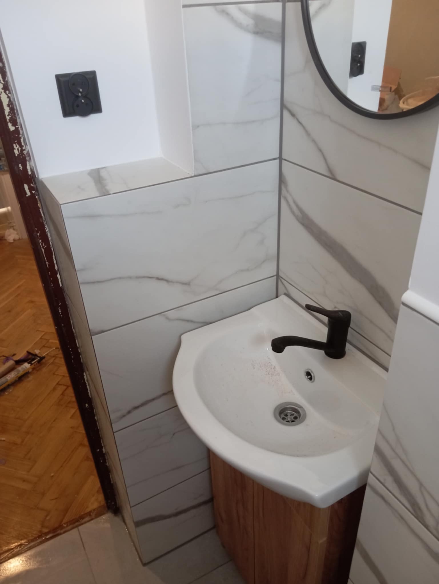 Umywalka z czarną armaturą na tle białych płytek imitujących marmur. Widoczne gniazdko elektryczne i fragment lustra. Wykończenie łazienki w nowoczesnym stylu.