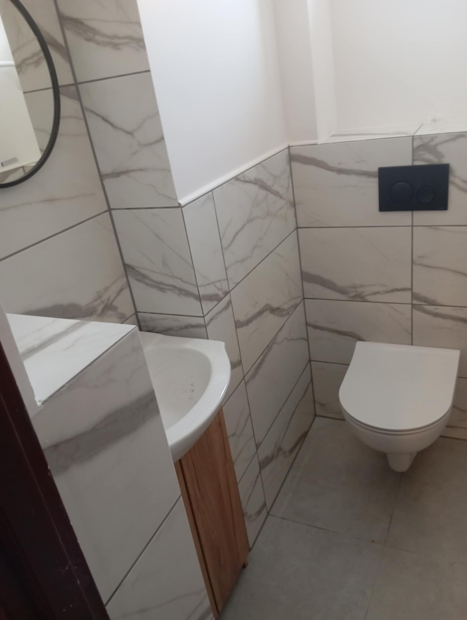 Wykończona toaleta z umywalką na szafce i podwieszaną miską WC. Białe płytki imitujące marmur na ścianach i podłodze. Okrągłe lustro.