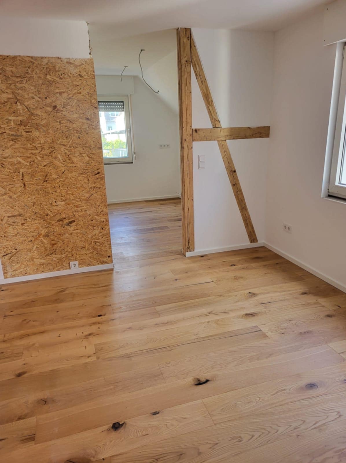 Pokój w trakcie remontu: jasne panele podłogowe, fragment ściany z płyt OSB, drewniana konstrukcja nośna w przejściu, okno w tle. Widoczne gniazdka elektryczne.