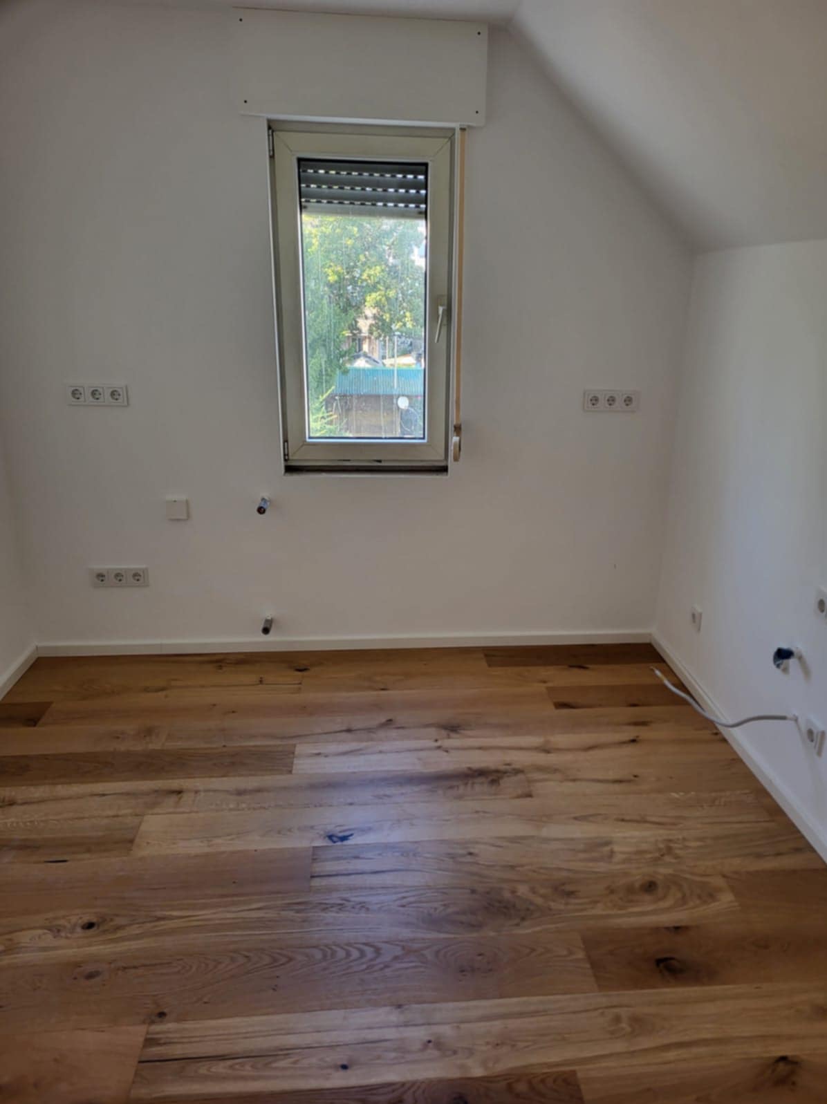 Jasny pokój po remoncie: świeżo położona drewniana podłoga, białe ściany z gniazdkami, okno z roletami. Widok na zieleń za oknem. Skosy sufitu.