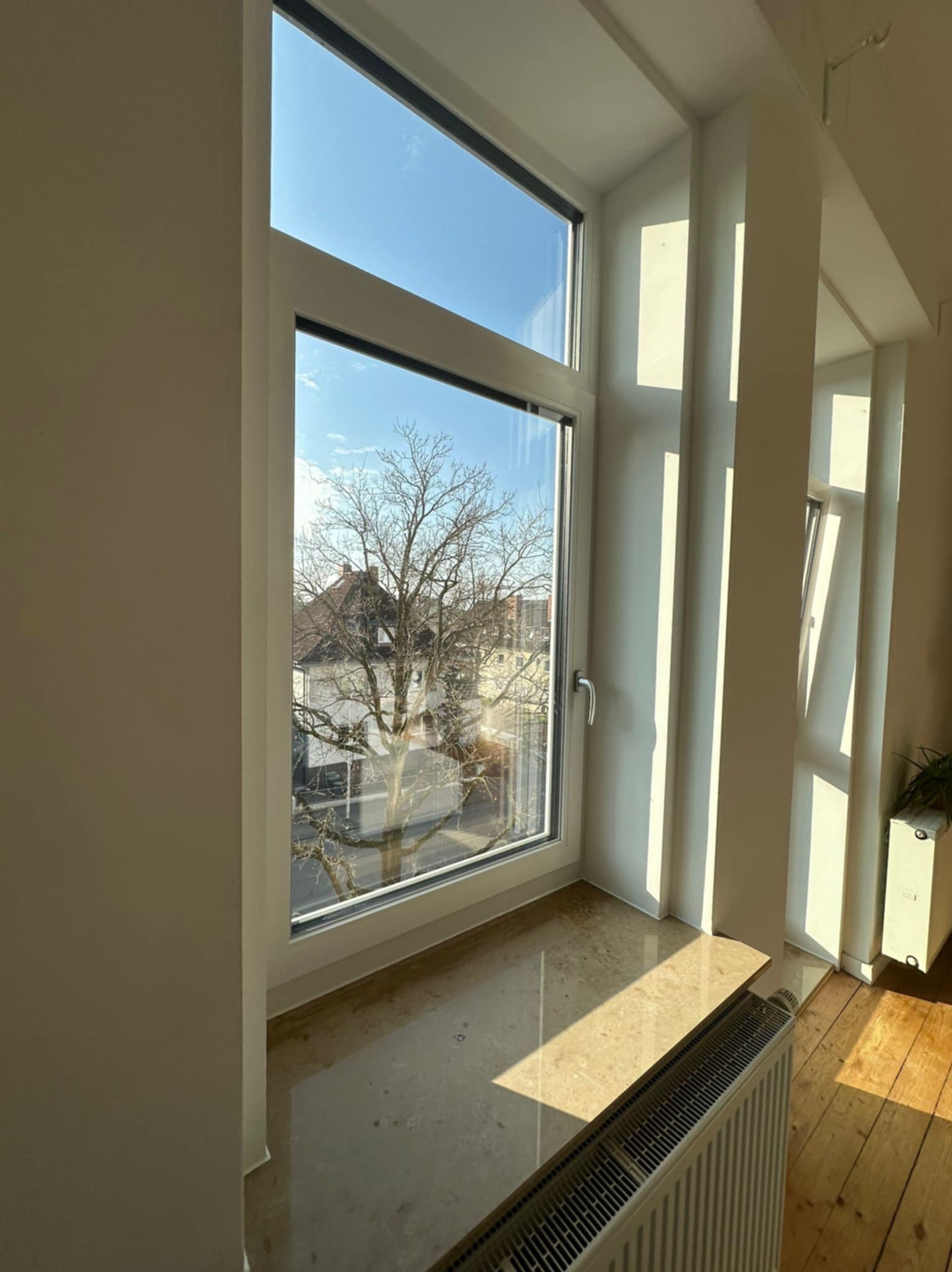 Jasny, beżowy parapet z marmuru pod oknem. Widok na drzewo i budynki. Grzejnik pod parapetem. Promienie słoneczne na podłodze.