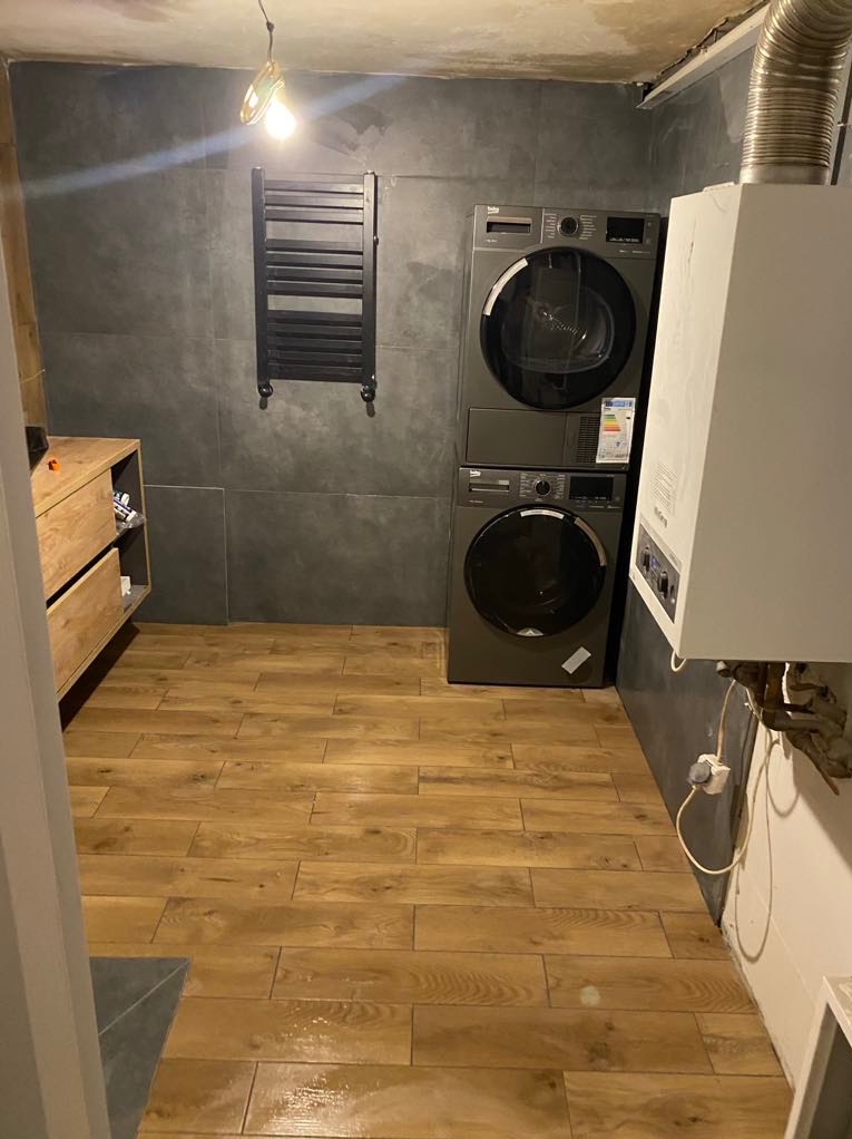Wykończona łazienka z drewnopodobną podłogą, szarymi płytkami ściennymi, grzejnikiem drabinkowym, pralko-suszarką w zabudowie i piecem gazowym.