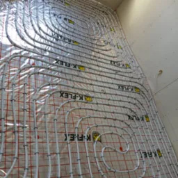 Rozłożone rury ogrzewania podłogowego K-FLEX na folii z siatką, przymocowane plastikowymi uchwytami, widok z góry pod kątem. Białe ściany w tle.