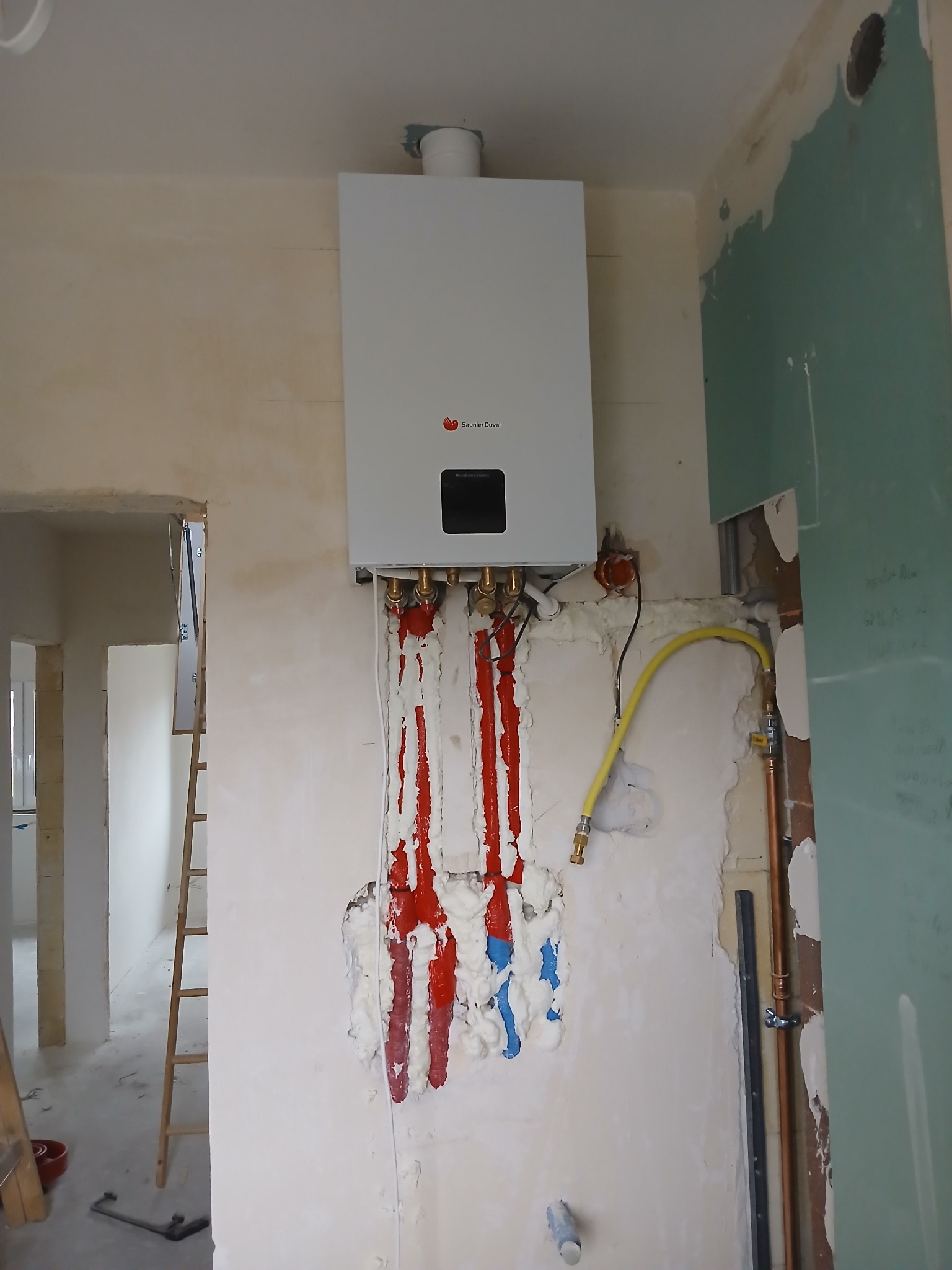 Montaż wiszącego kotła gazowego Saunier Duval na ścianie w trakcie prac wykończeniowych, widoczne rury w izolacji piankowej, żółty przewód gazowy i fragment drabiny w tle.