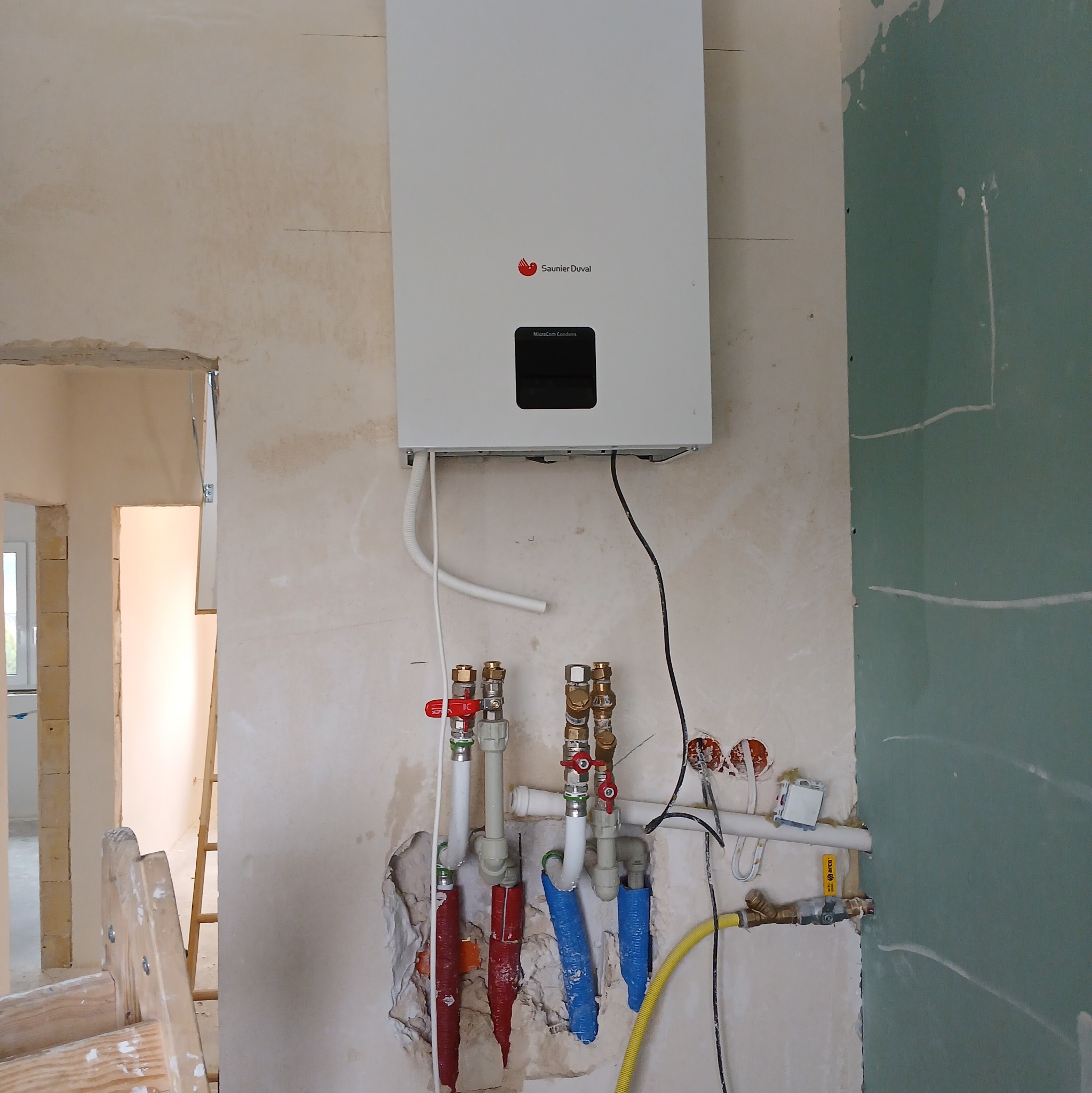 Biały wiszący piec gazowy Saunier Duval z podłączeniami hydraulicznymi i gazowymi w surowym wnętrzu, widoczne rury z zaworami, instalacja elektryczna i fragment ściany z płyt gipsowo-kartonowych.