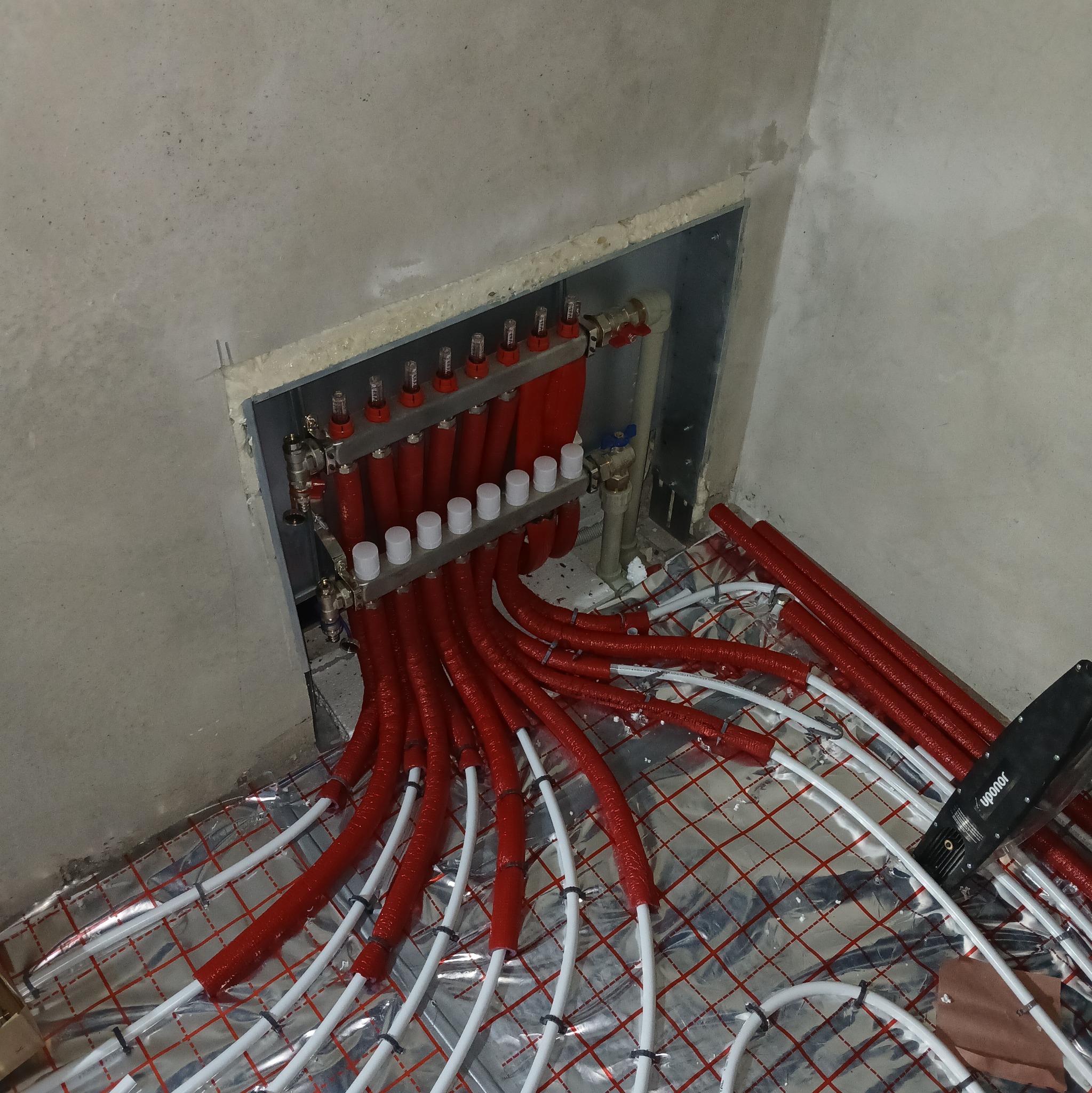 Rozdzielacz ogrzewania podłogowego z czerwonymi rurami w izolacji, ułożonymi na folii refleksyjnej z siatką, widoczna zaciskarka Uponor.