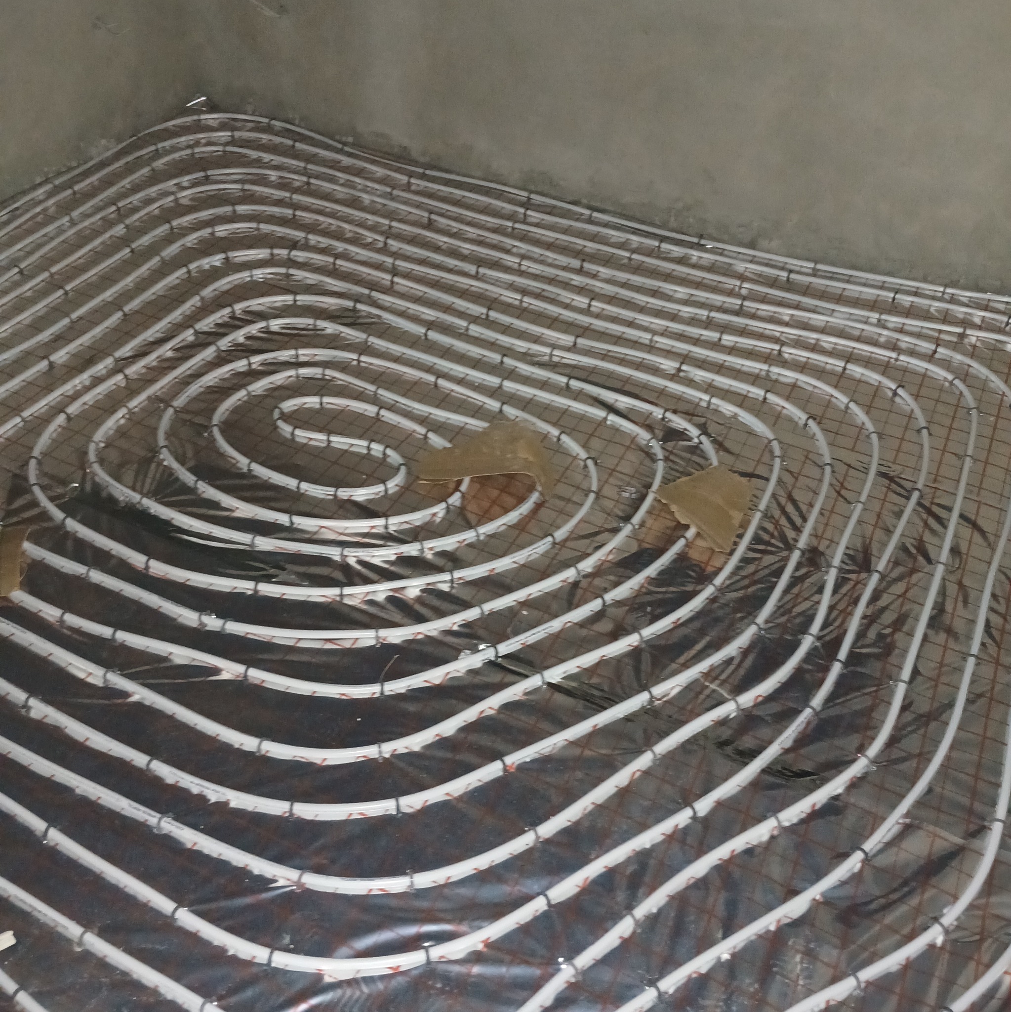 Białe rury ogrzewania podłogowego ułożone spiralnie na folii izolacyjnej, przymocowane do metalowej siatki, widok z góry, surowy stan przed wylewką.