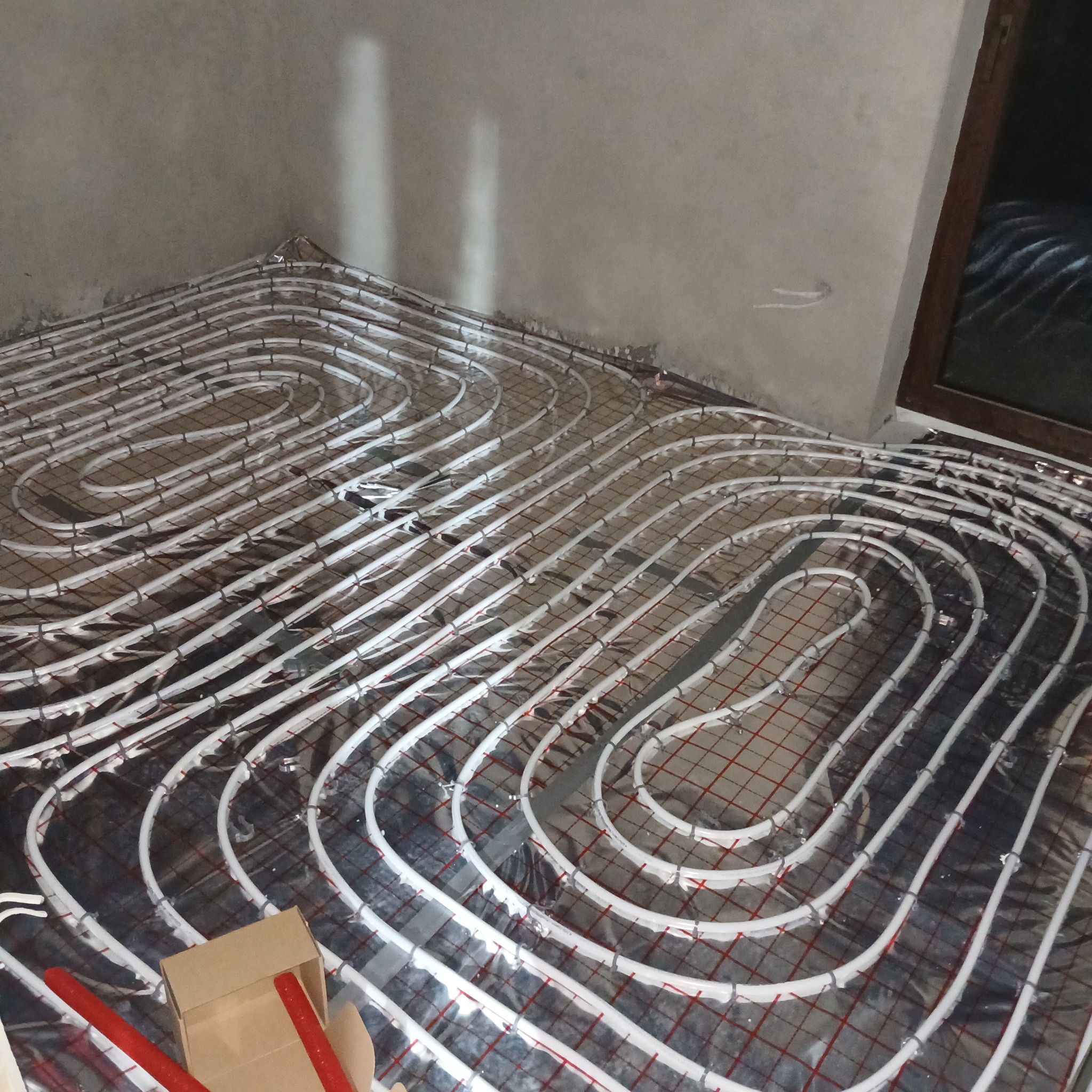 Ułożone białe rury ogrzewania podłogowego na folii aluminiowej z siatką, widoczne pudełko i czerwone elementy montażowe.
