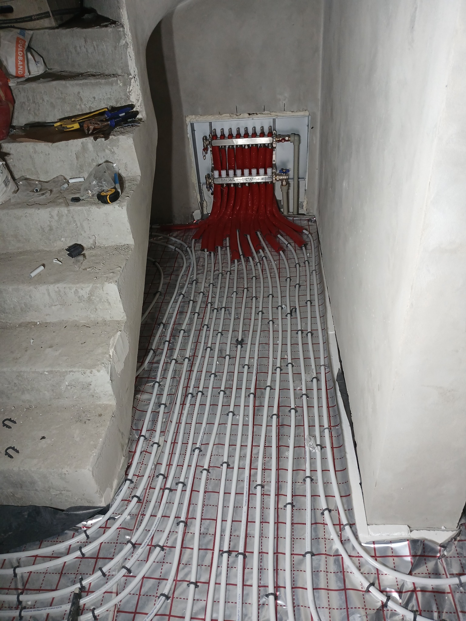 Rozłożona instalacja ogrzewania podłogowego z czerwonymi rurami przy rozdzielaczu, widoczne narzędzia na schodach obok, Ostrołęka.