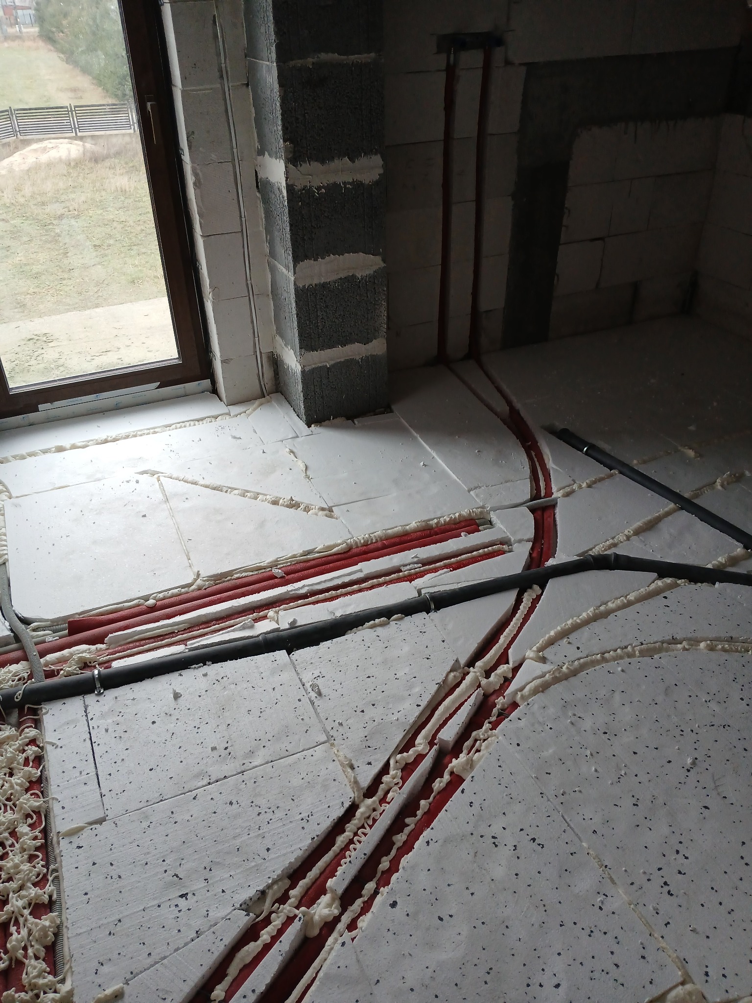 Podłoga w trakcie montażu ogrzewania podłogowego: czerwone rury w wycięciach styropianu, uszczelnione pianką montażową, widok na okno i surową ścianę z bloczków.