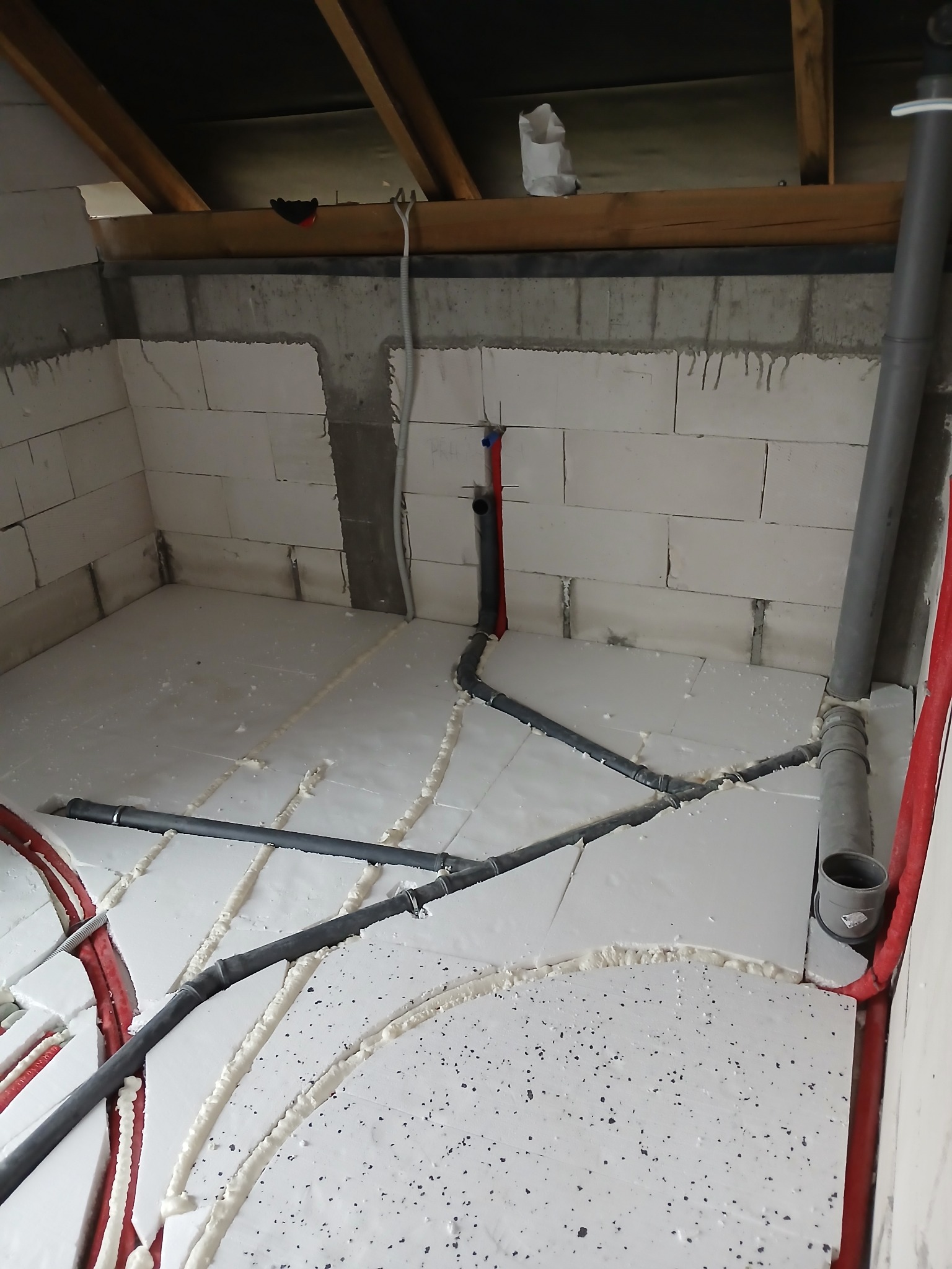 Wnętrze w trakcie prac instalacyjnych: rury grzewcze i kanalizacyjne ułożone na białej izolacji podłogowej, ściany z białych bloczków, widoczna konstrukcja dachu.