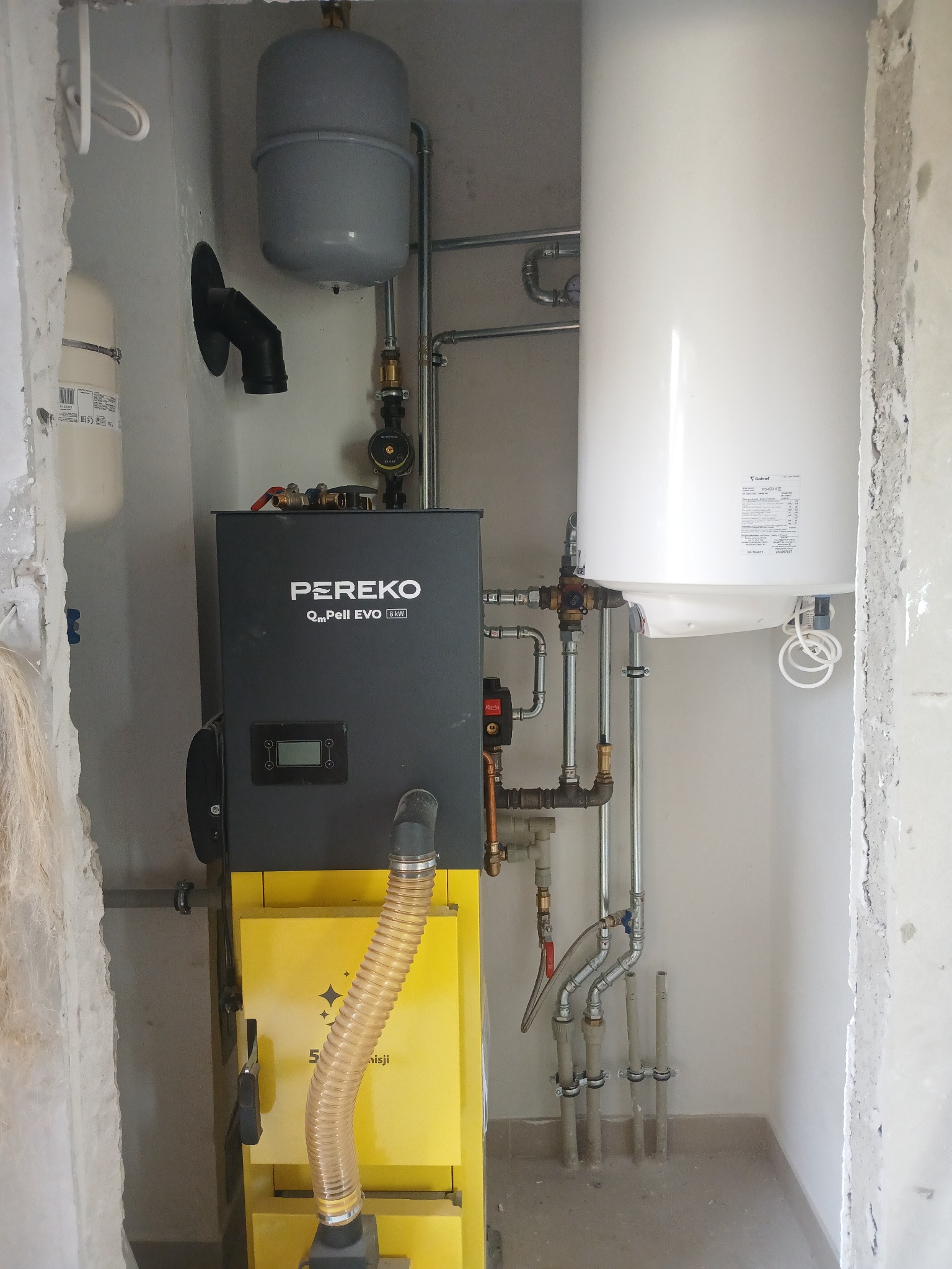 Kompaktowa kotłownia: czarny piec Pereko 8kW z żółtym zasobnikiem pelletu i białym zasobnikiem CWU 100l, rury instalacji w małym pomieszczeniu o powierzchni 1.5m2.