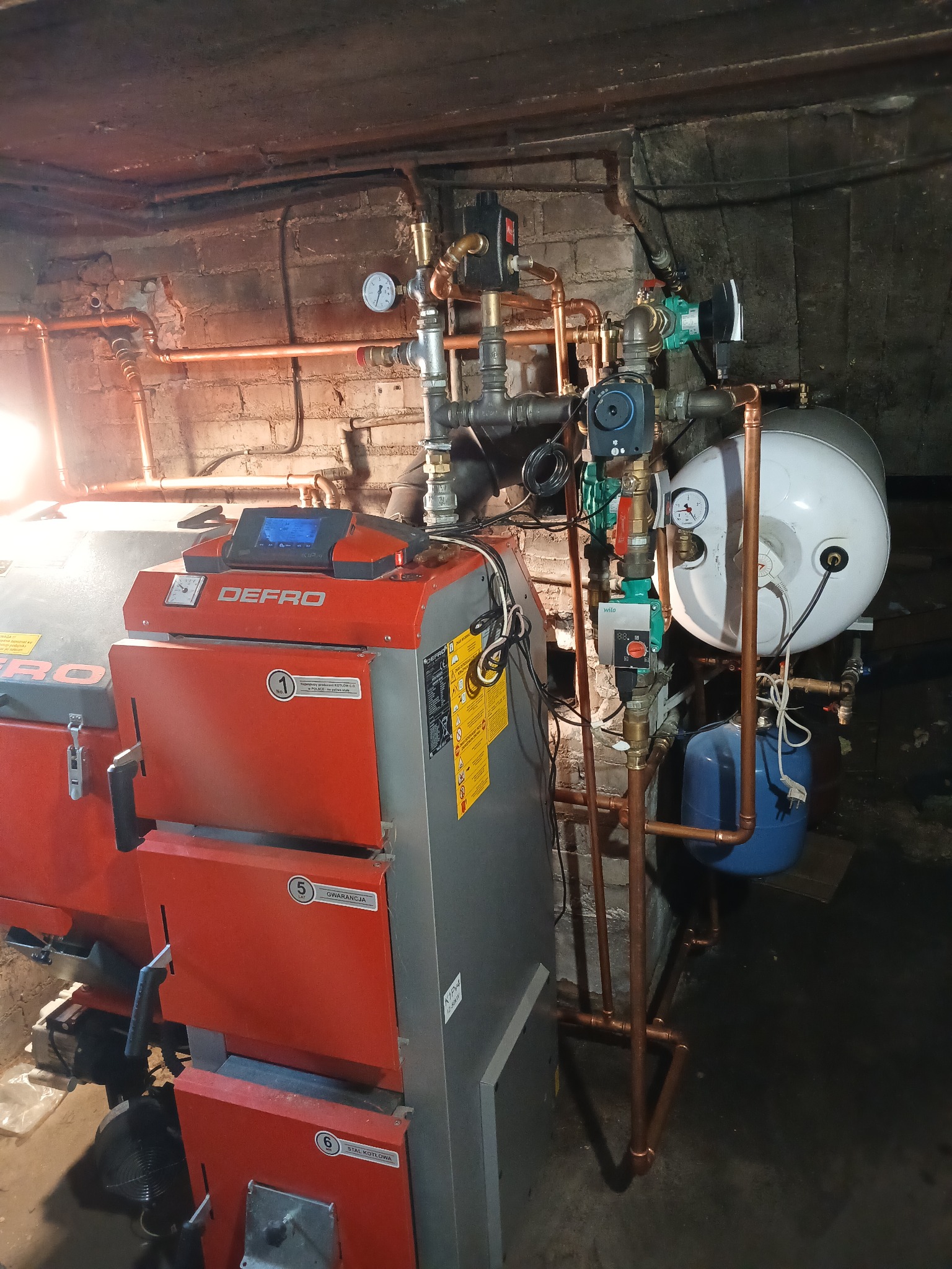 Kotłownia z piecem Defro, instalacją CO z miedzianych rur, zbiornikiem CWU i pompą obiegową. Manometry i zawory na instalacji. Surowa ściana z cegły w tle.