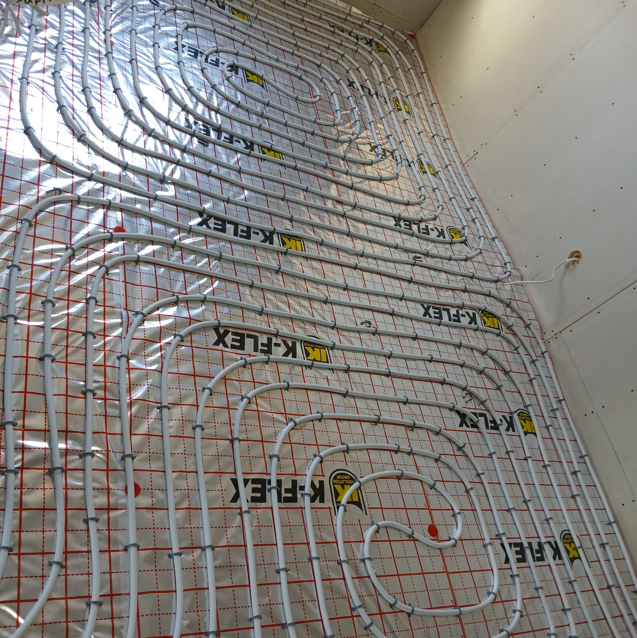 Rozłożone rury ogrzewania podłogowego K-FLEX na folii z siatką, przymocowane plastikowymi uchwytami, widok z góry pod kątem. Białe ściany w tle.