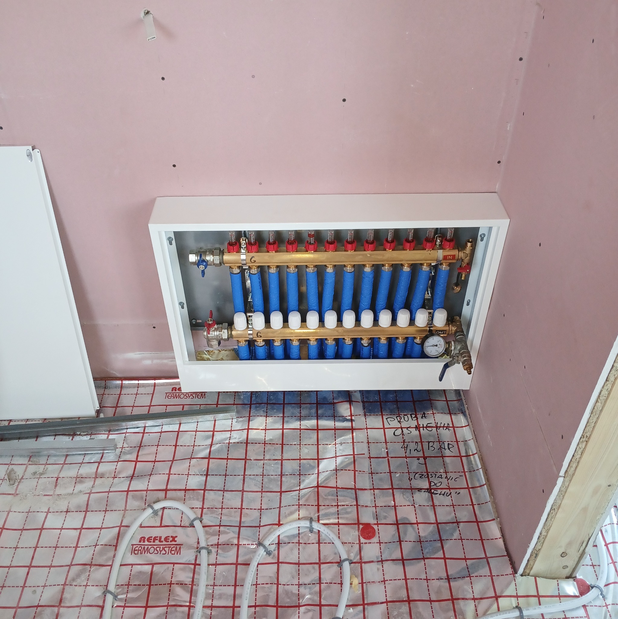 Rozdzielacz ogrzewania podłogowego w białej obudowie, widoczne rury w izolacji i manometr. Na podłodze mata grzewcza z zapiskami, ściany w surowym stanie.