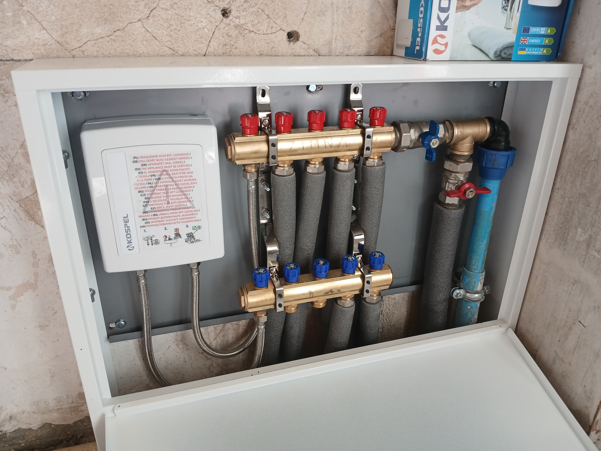 Rozdzielacz ogrzewania podłogowego Kospel w szafce, z widocznymi rurami w izolacji, zaworami z czerwonymi i niebieskimi pokrętłami oraz elementami instalacji.