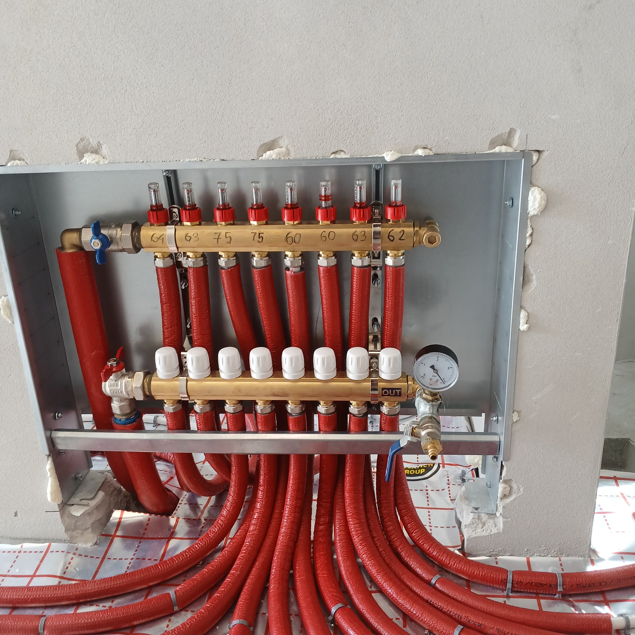 Rozdzielacz ogrzewania podłogowego z czerwonymi rurami, zamontowany na ścianie z widoczną izolacją. Manometr i zawory regulacyjne. Instalacja w trakcie montażu.