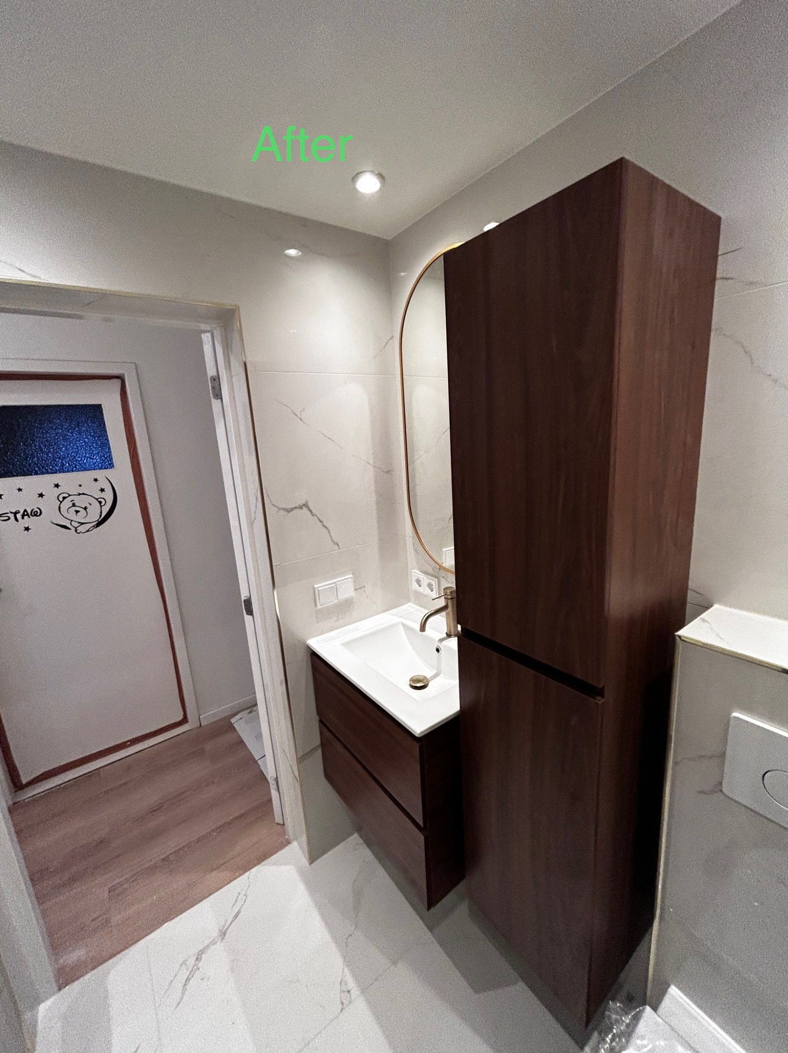 Wykończona łazienka z marmurowymi płytkami, brązową szafką pod umywalką, wysoką szafką wiszącą i okrągłym lustrem z podświetleniem, widok na drzwi z naklejką misia.