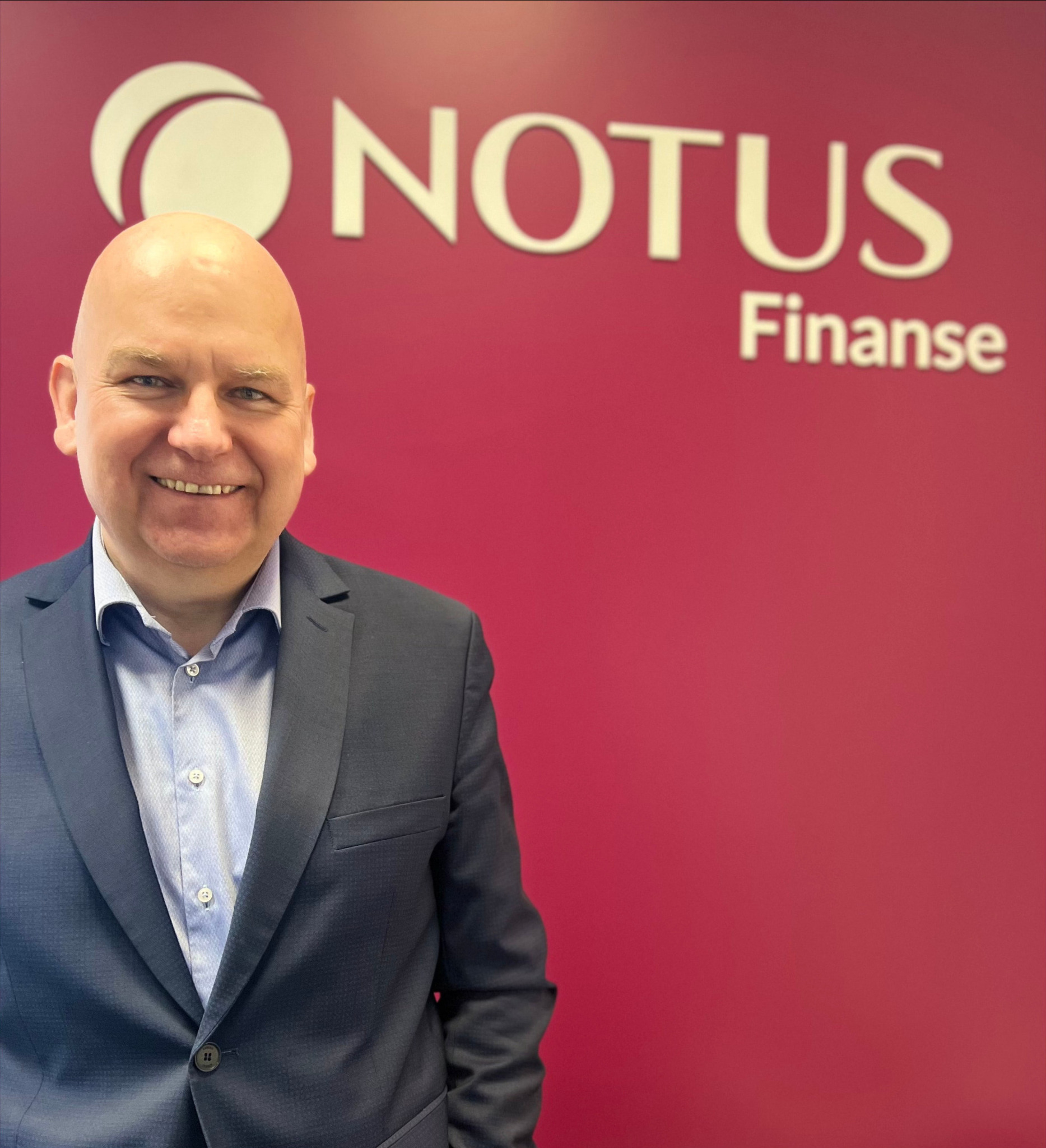 Uśmiechnięty mężczyzna w garniturze na tle ściany z logo firmy finansowej.