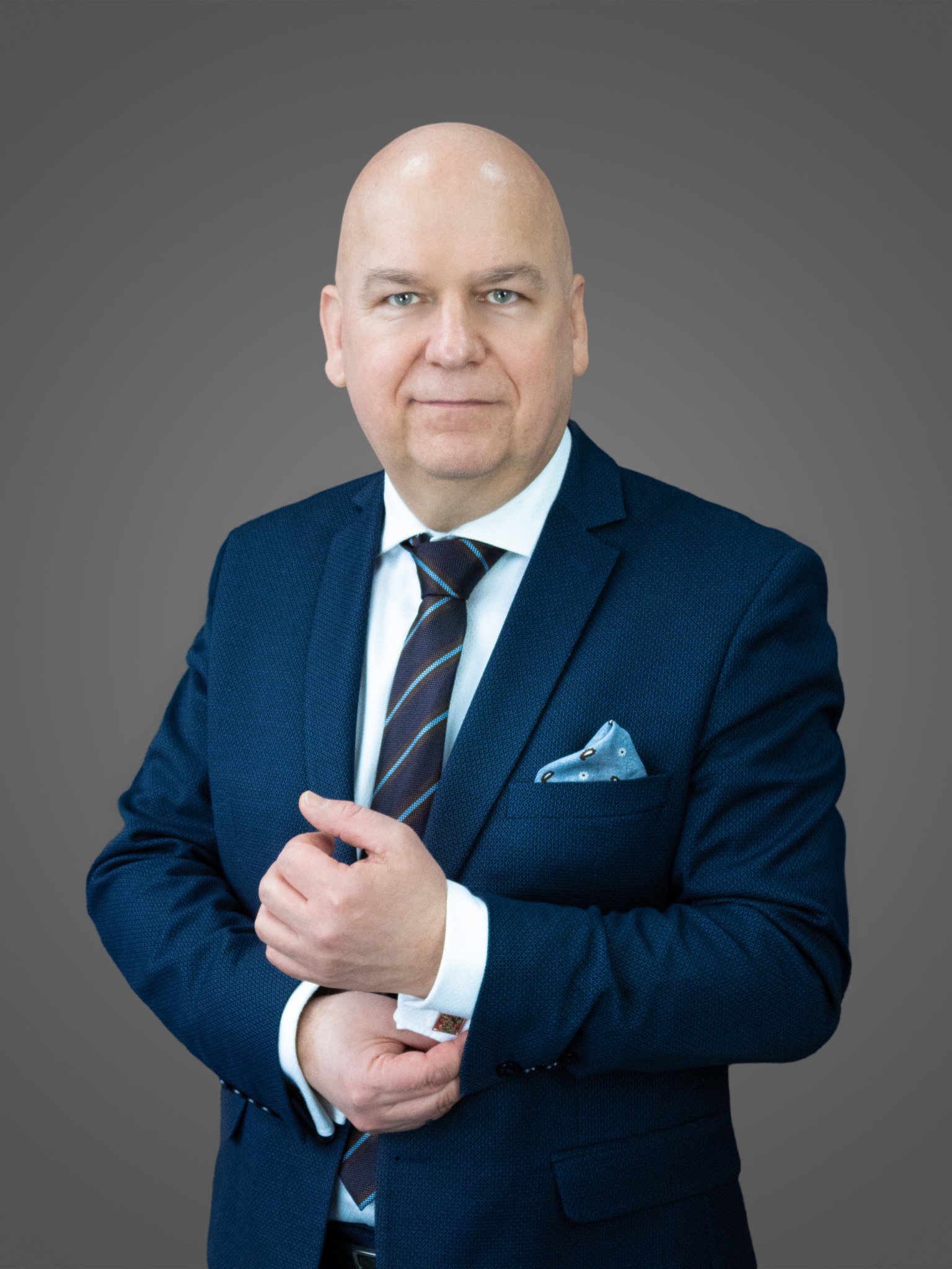 Portret mężczyzny w granatowym garniturze, z chusteczką w kieszonce i mankietami z ozdobnymi spinkami, na jednolitym szarym tle.