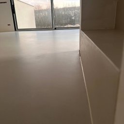 Mateoprojekt - Jasna, jednolita posadzka w nowym pomieszczeniu z dużym oknem, widoczne detale wykończenia przy ścianie i progu.