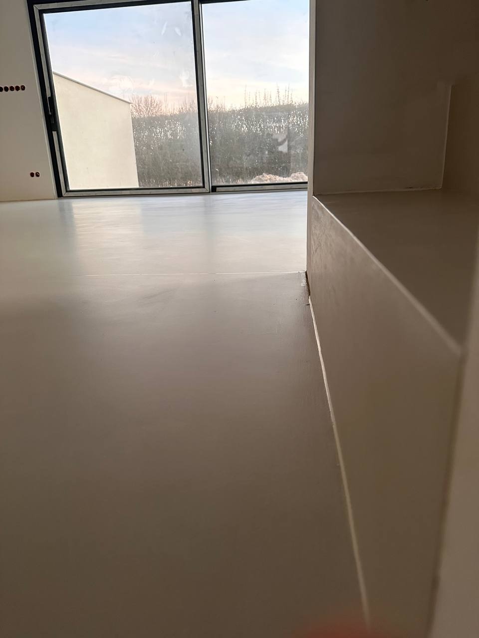 Jasna, jednolita posadzka w nowym pomieszczeniu z dużym oknem, widoczne detale wykończenia przy ścianie i progu.