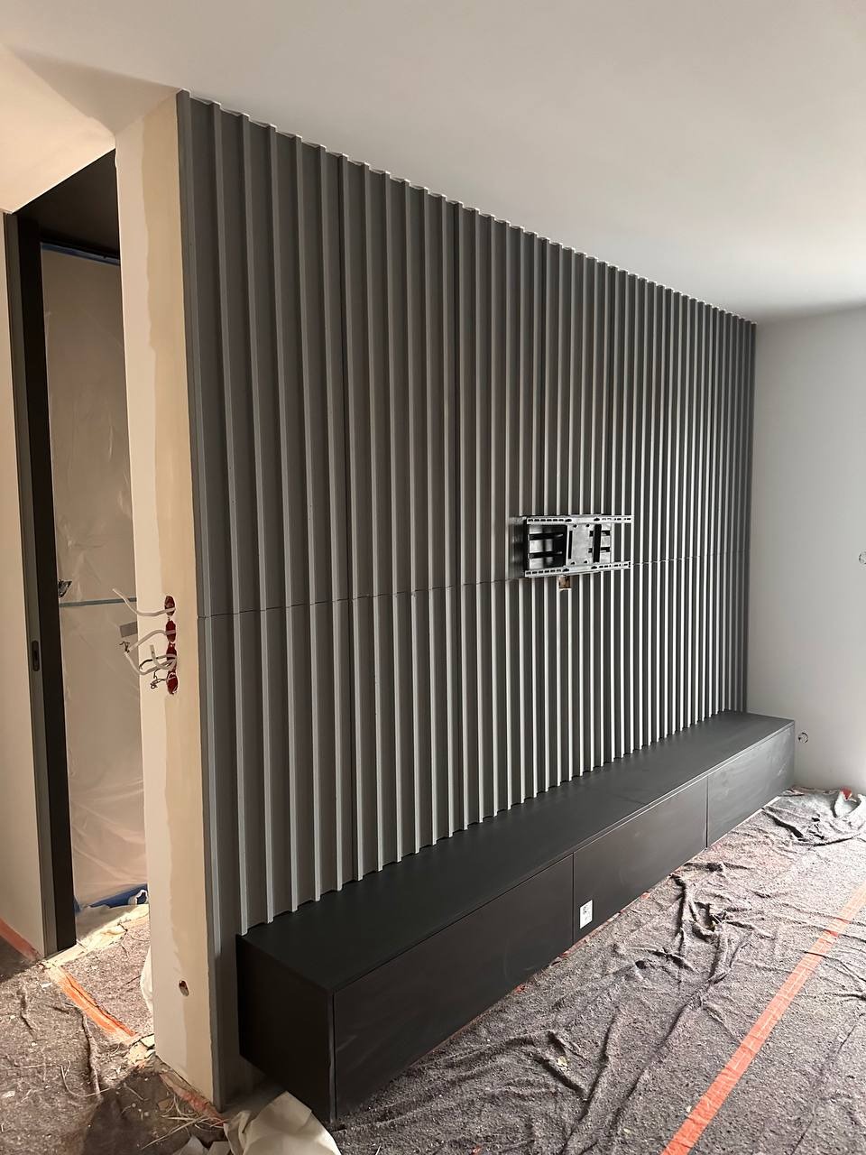 Ściana z szarych paneli dekoracyjnych z zamontowanym uchwytem TV i czarną szafką RTV, zabezpieczona folią podłoga w trakcie prac wykończeniowych.