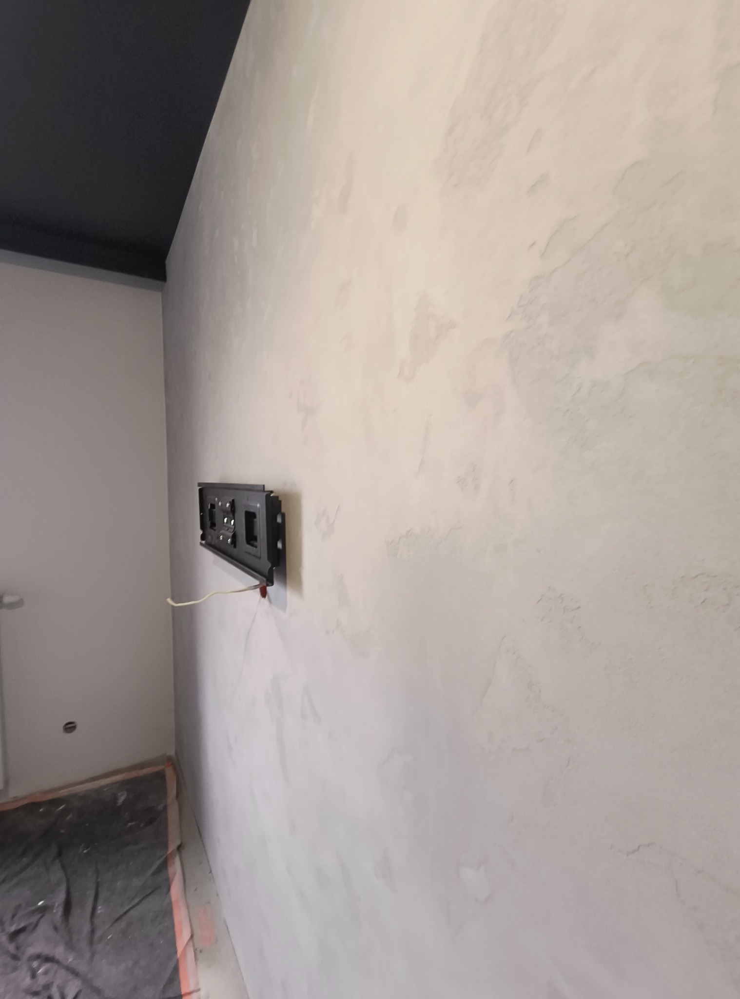 Ściana pokryta dekoracyjną, jasną masą z widoczną fakturą, zamontowany uchwyt pod telewizor, fragment czarnego sufitu podwieszanego, podłoga zabezpieczona folią malarską.
