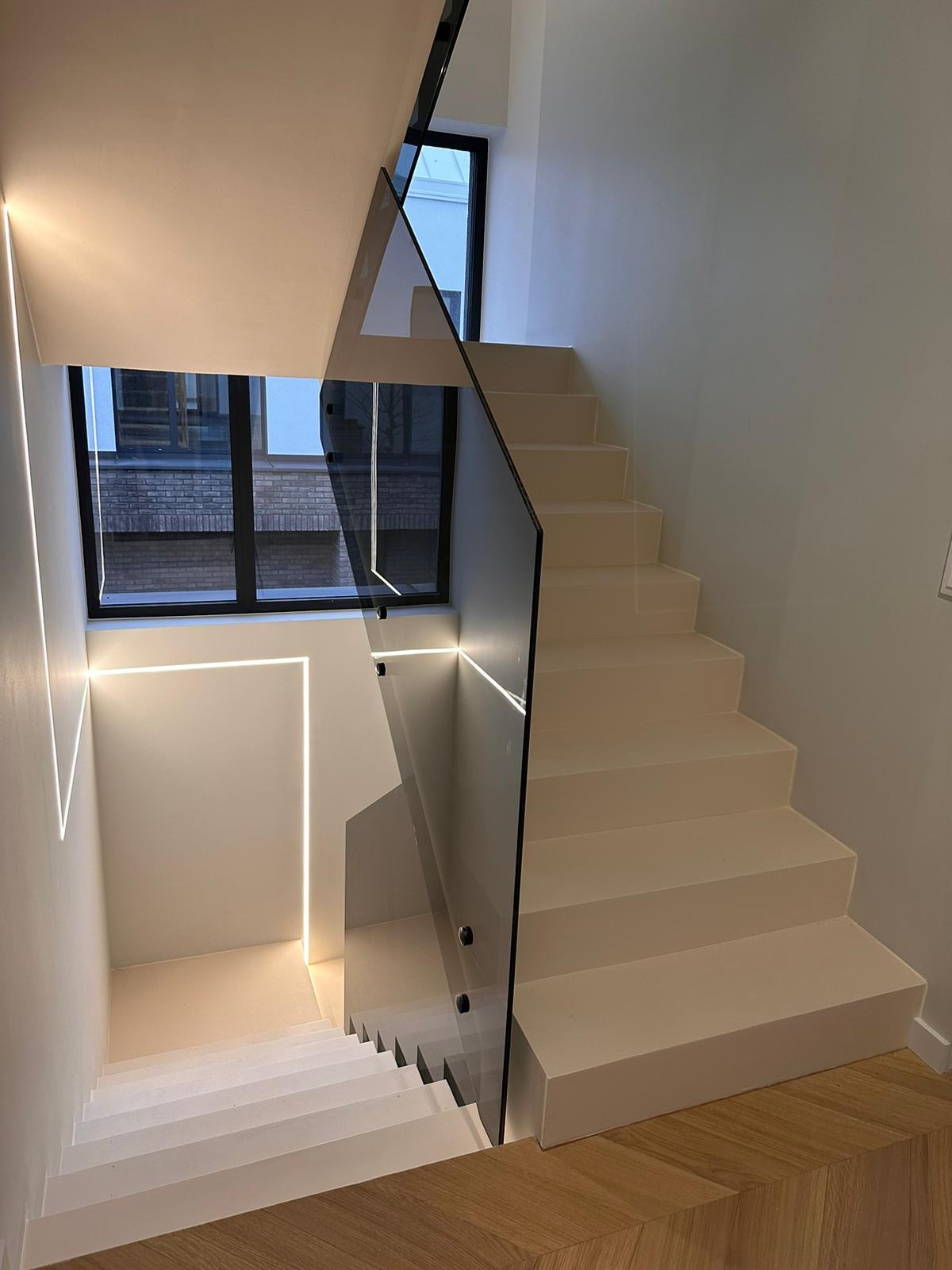 Nowoczesne schody z białymi stopniami i szklaną balustradą, oświetlone liniowymi lampami LED w ścianie i balustradzie, widoczne drewniane panele podłogowe.