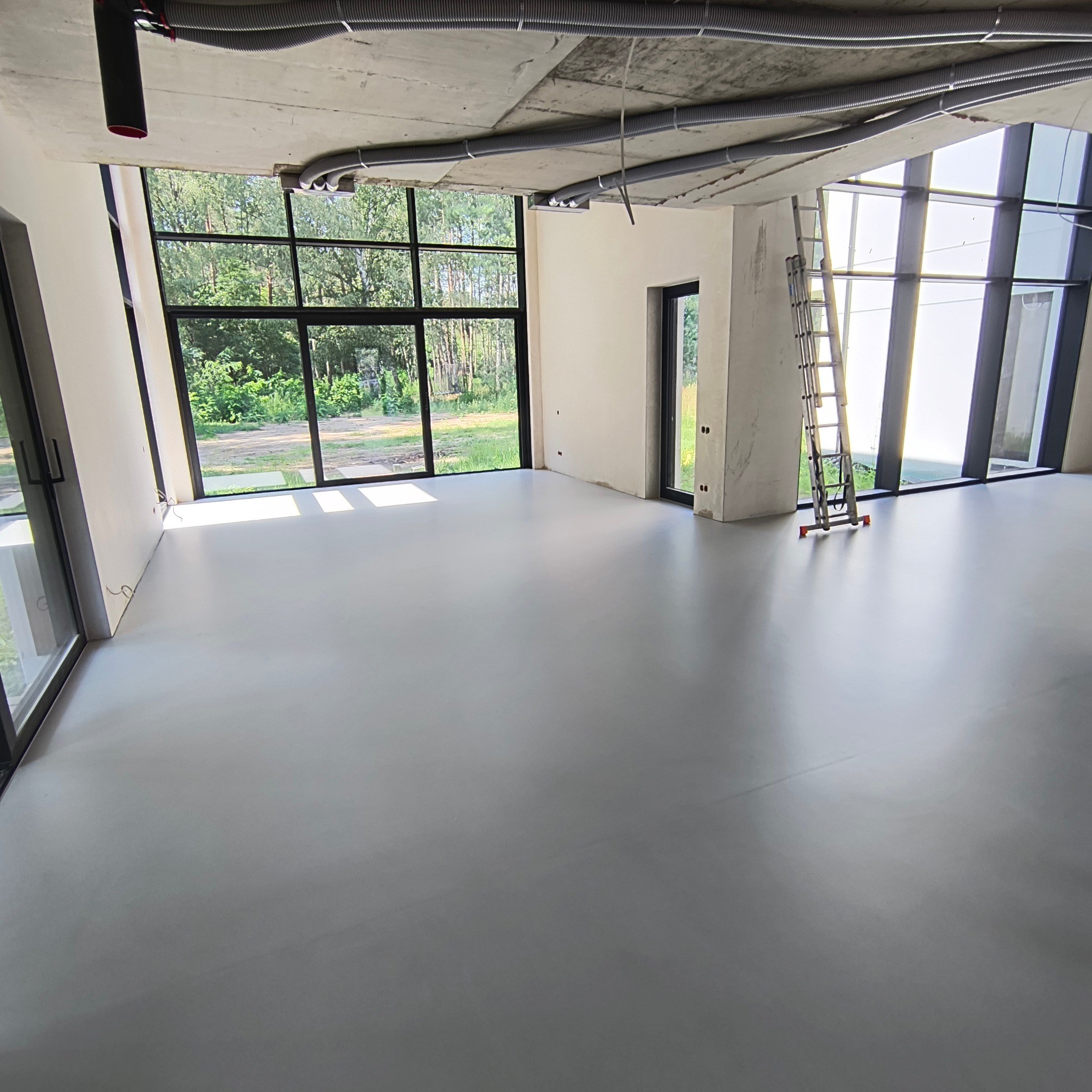 Jasnoszara, gładka posadzka w przestronnym, surowym wnętrzu z dużymi oknami wychodzącymi na zieleń, widoczne rury wentylacyjne pod betonowym stropem i aluminiowa drabina.