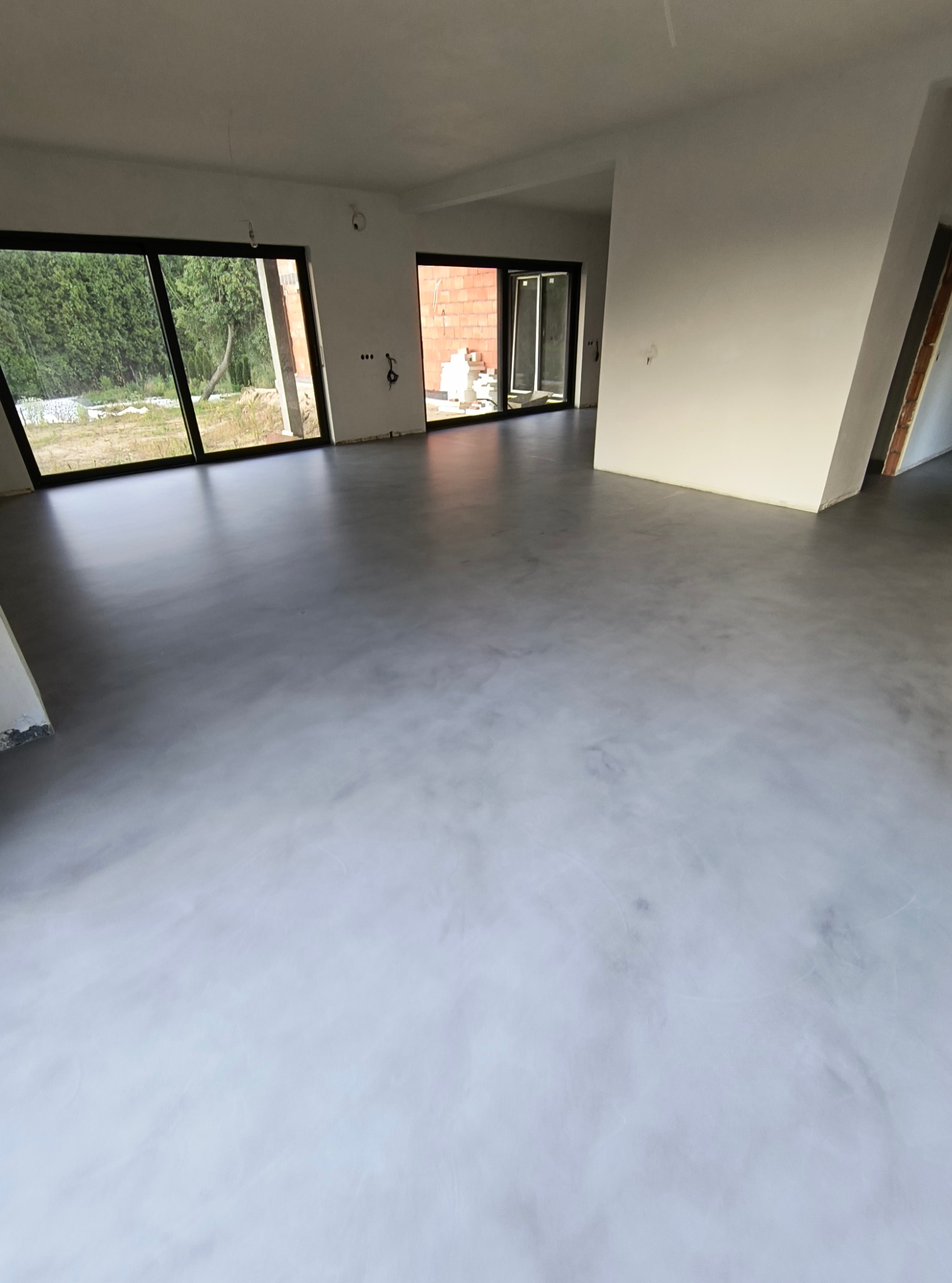 Wnętrze domu w trakcie wykańczania z położoną szarą, błyszczącą posadzką żywiczną, widoczne duże okna z czarnymi ramami i surowe ściany.