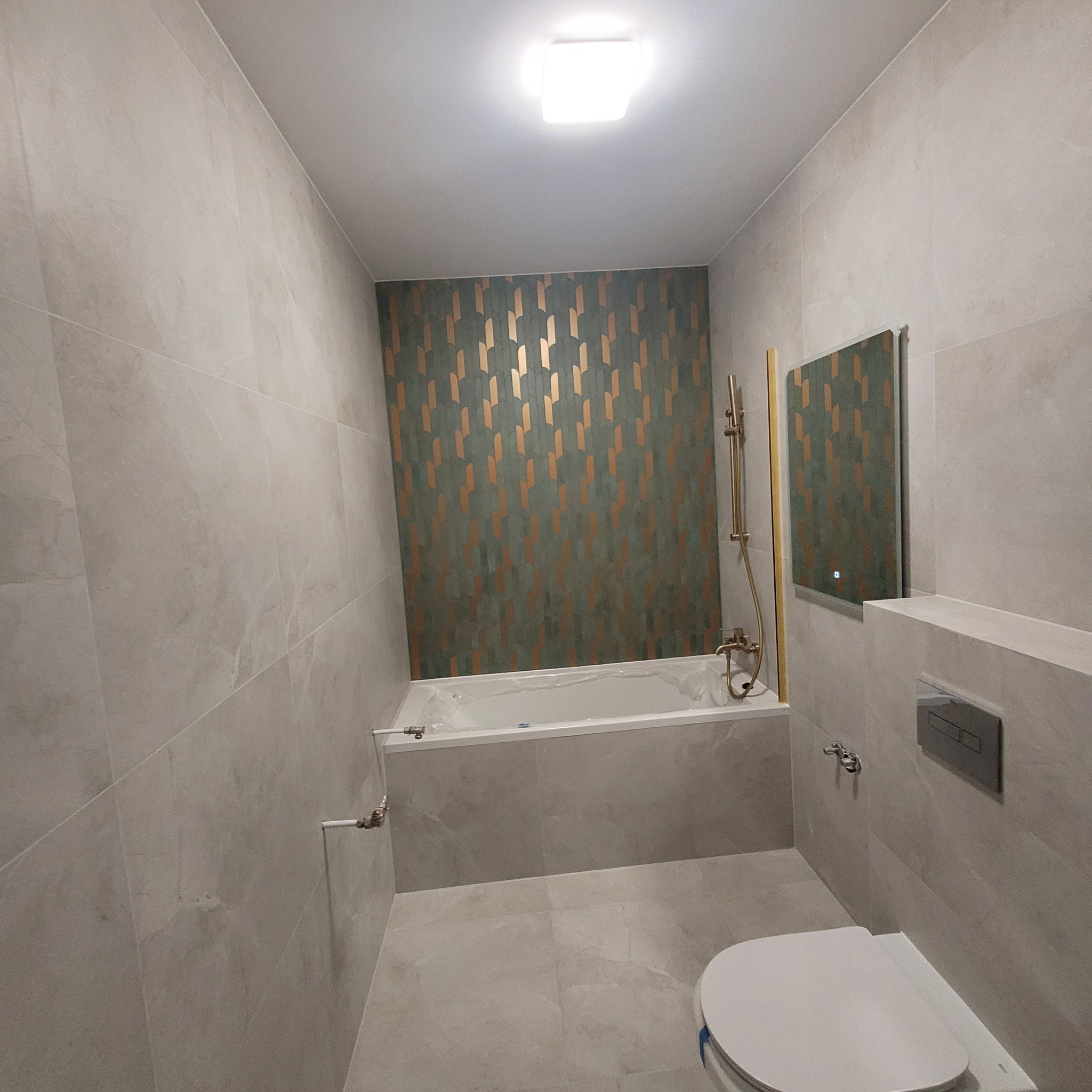 Nowoczesna łazienka z płytkami imitującymi marmur na ścianach i podłodze, akcentująca ściana z geometrycznym wzorem w odcieniach zieleni i złota, wanna częściowo zakryta folią ochronną, złota...