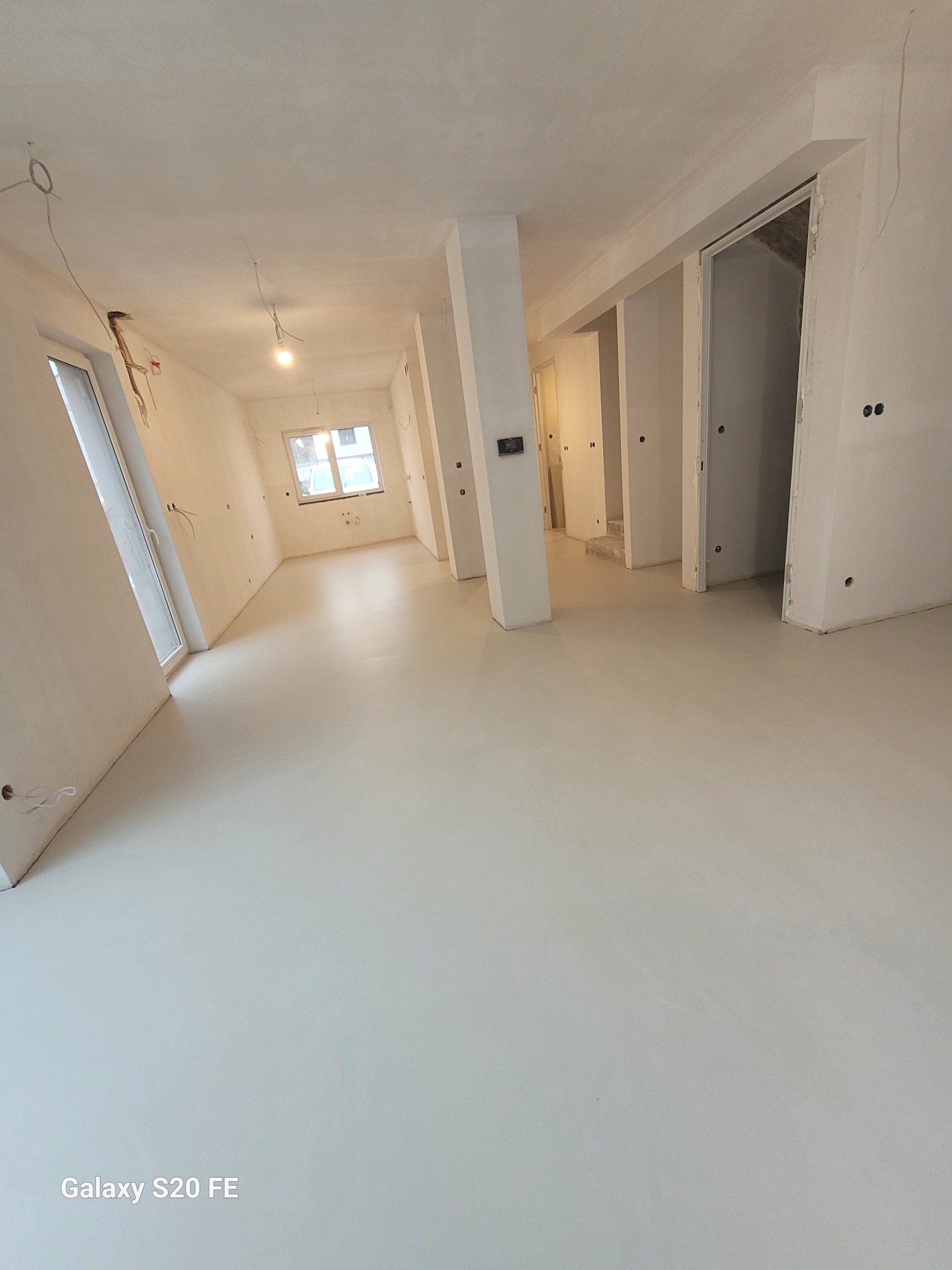 Jasne, przestronne wnętrze w trakcie prac wykończeniowych; widoczne ściany po nałożeniu warstwy wykończeniowej, gładka, jasna posadzka, oraz instalacje elektryczne wystające ze ścian.