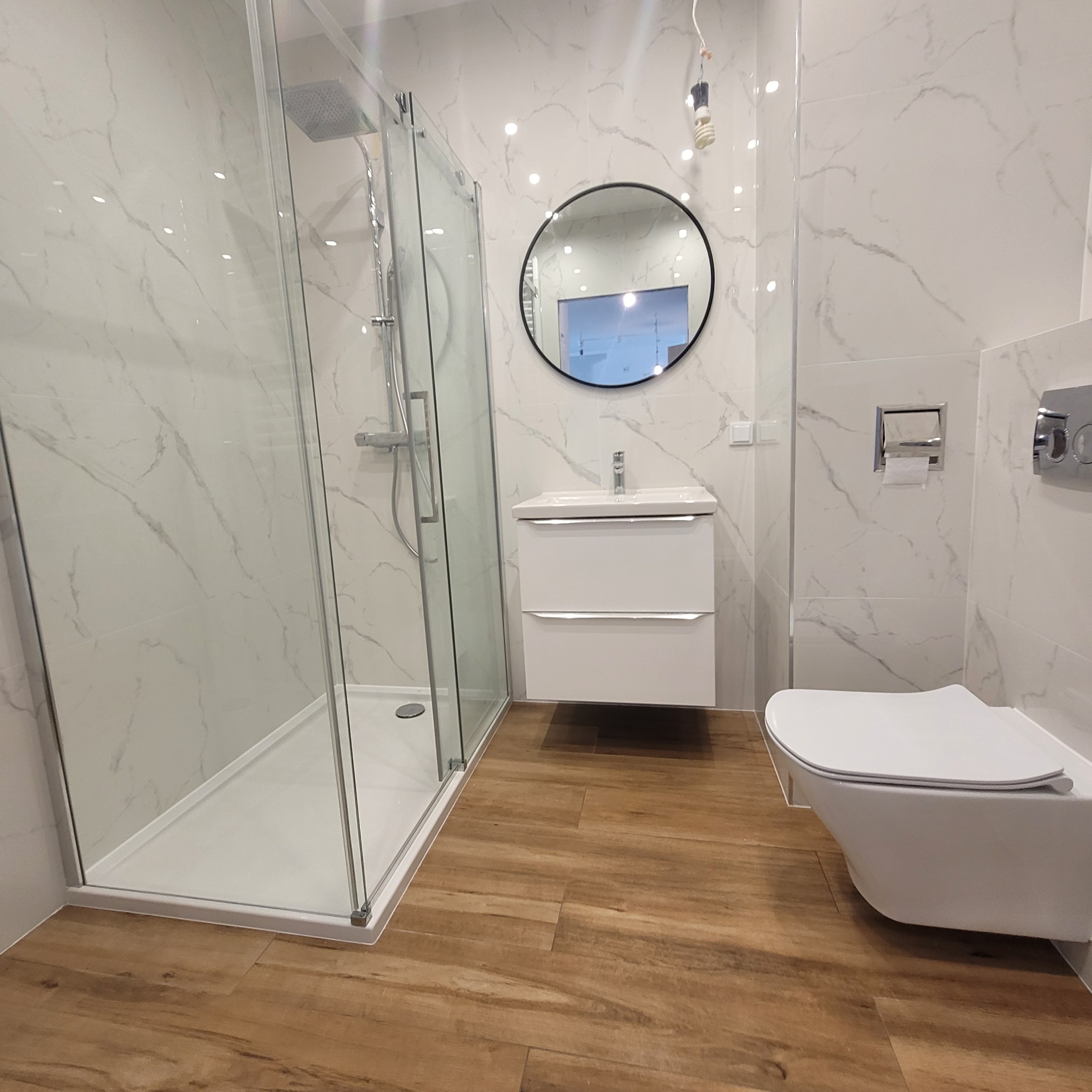 Nowoczesna łazienka po remoncie: kabina prysznicowa typu walk-in z deszczownicą, okrągłe lustro nad białą szafką z umywalką, podwieszana toaleta i drewnopodobna podłoga.