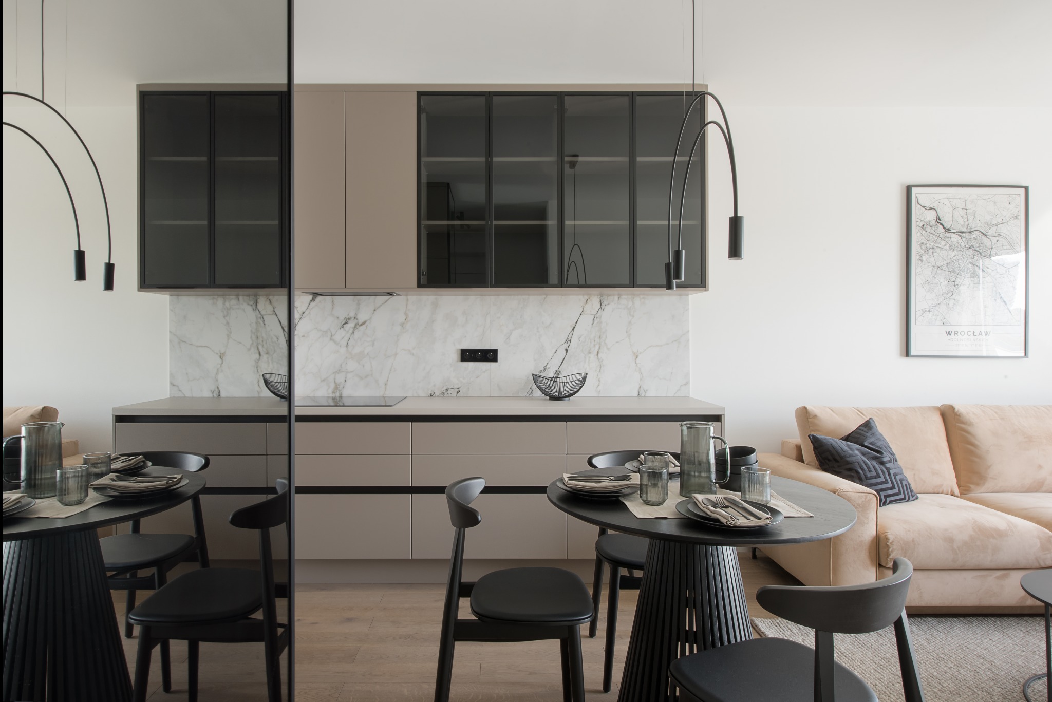 Nowoczesna kuchnia z beżowymi szafkami, marmurowym backsplashiem i czarnymi detalami, w tle salon z beżową sofą i plakatem z mapą Wrocławia.