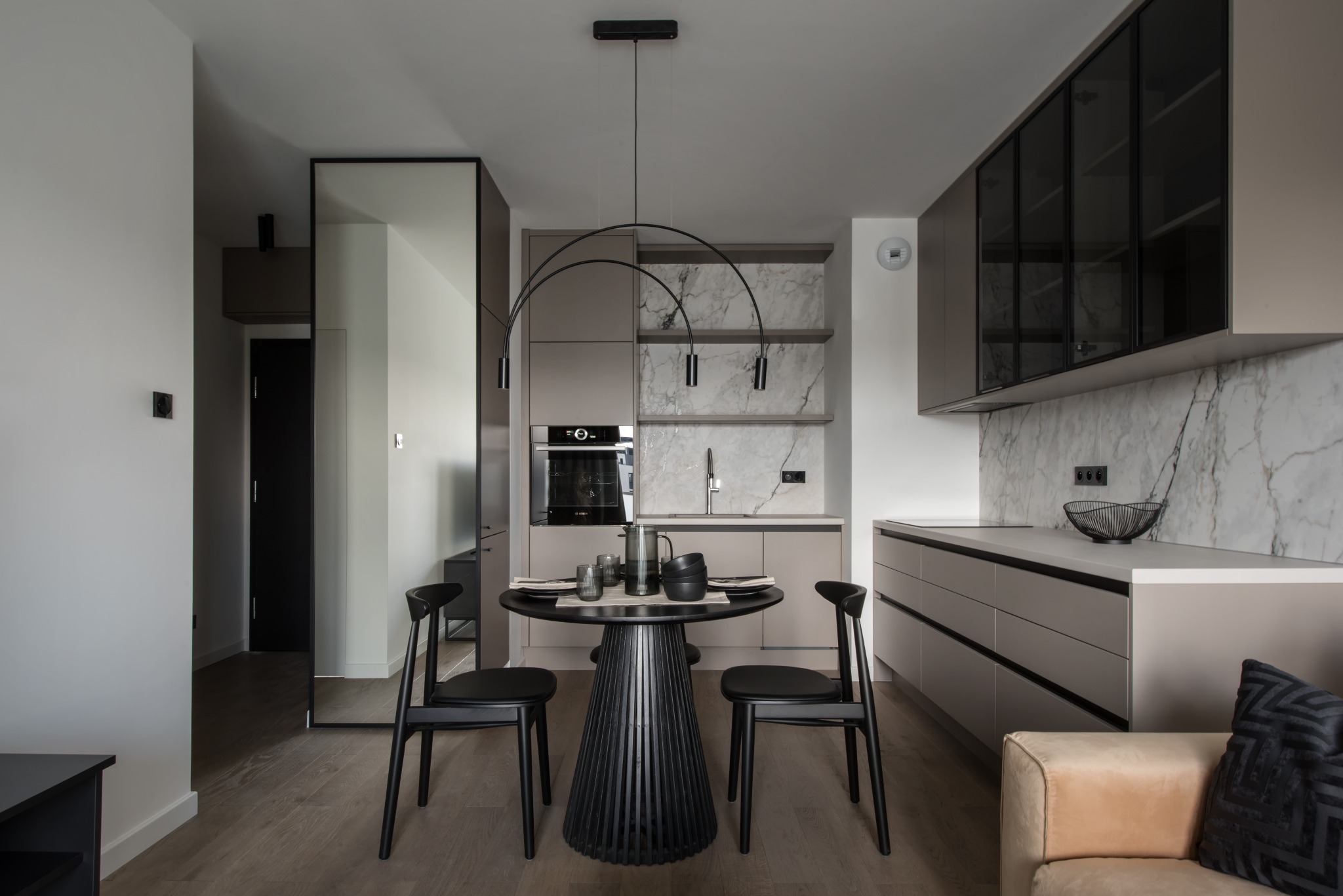 Nowoczesna kuchnia z zabudową w odcieniach beżu i marmuru, okrągły stół z czarnymi krzesłami i designerską lampą łukową.
