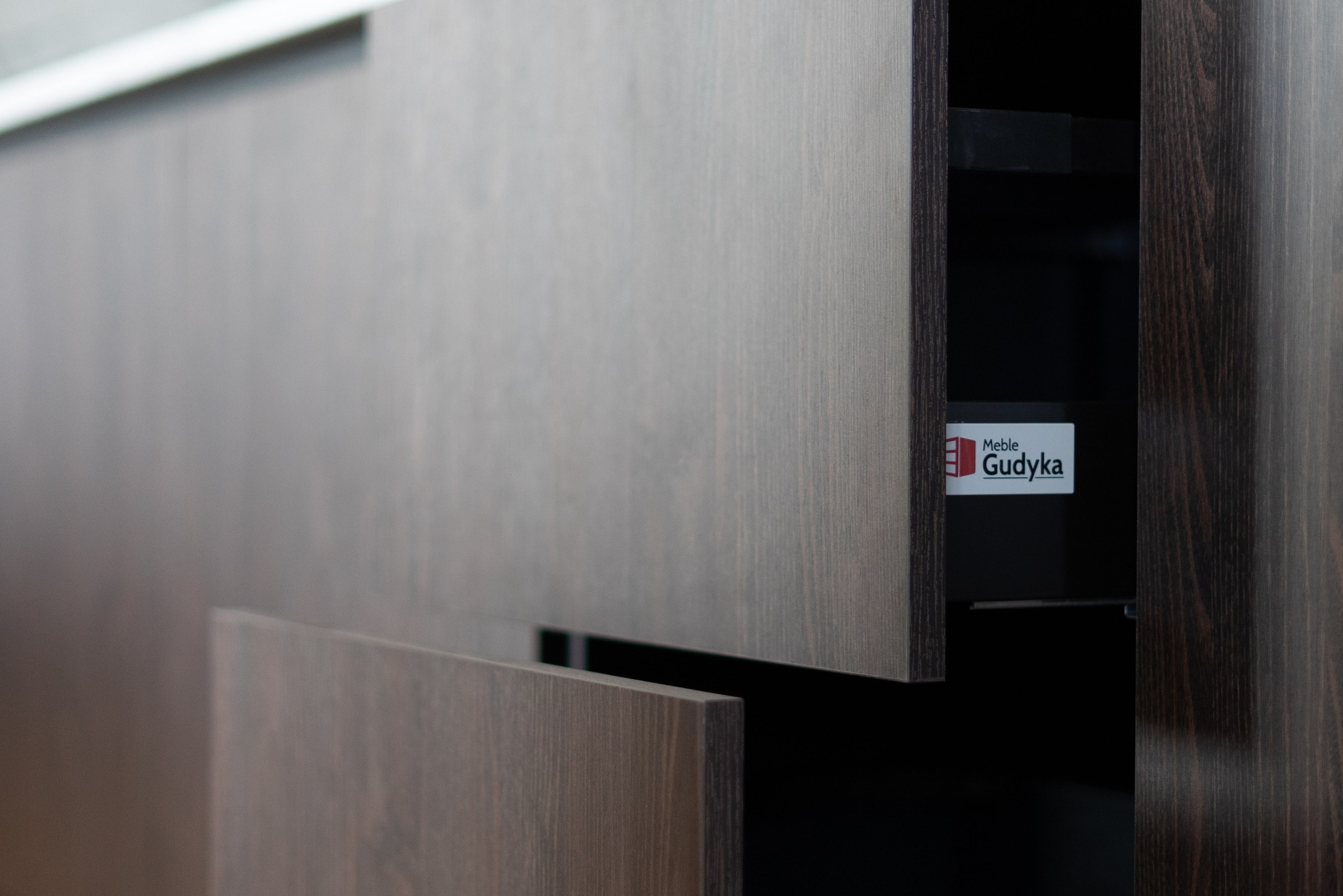 Ciemnobrązowe, drewniane fronty szuflad kuchennych z widocznym logotypem producenta Meble Gudyka wewnątrz otwartej szuflady.