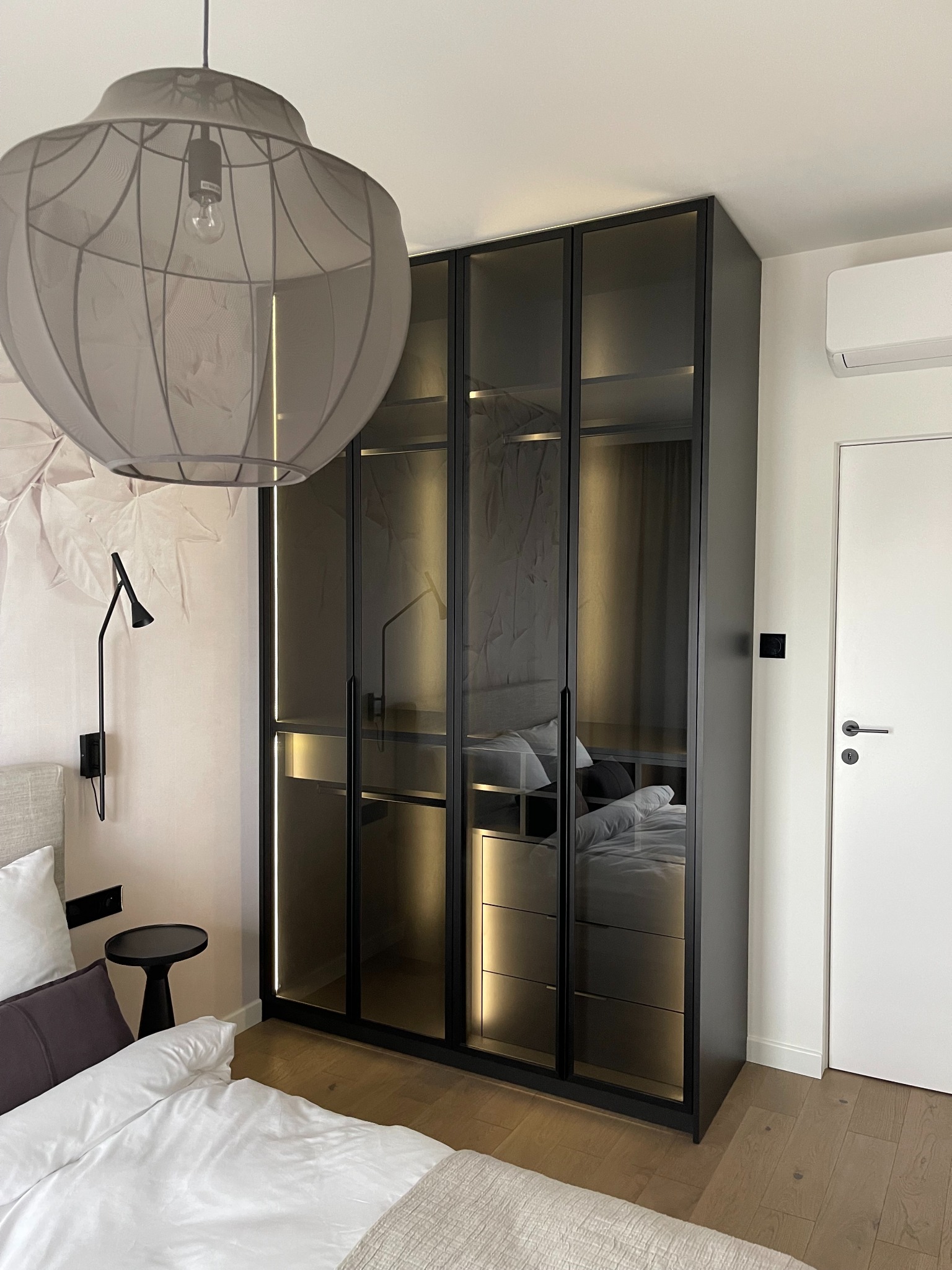 Czarna szafa wnękowa z przesuwnymi, szklanymi drzwiami, odbijająca wnętrze sypialni, z widocznymi półkami i szufladami, oświetlona listwami LED, obok białe drzwi i łóżko.