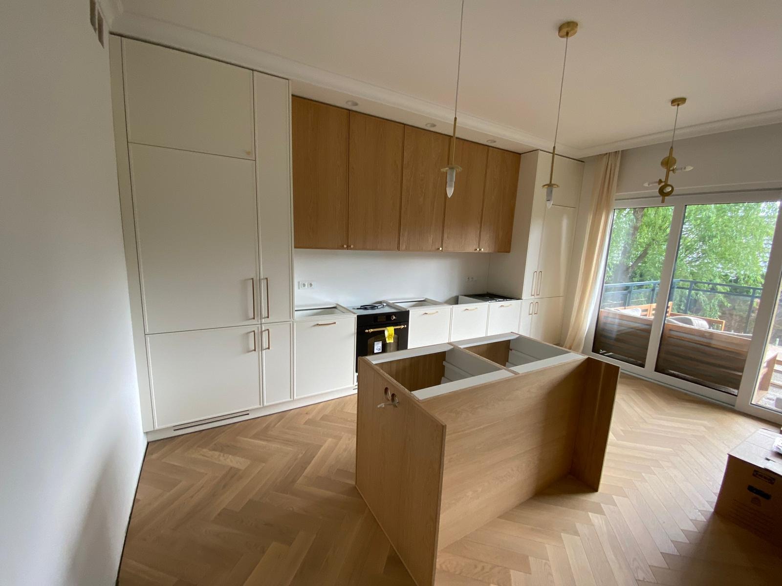 Wnętrze kuchni z drewnianą podłogą w jodełkę, białe i drewniane szafki kuchenne, wyspa kuchenna w trakcie montażu, widok na taras przez duże okno.