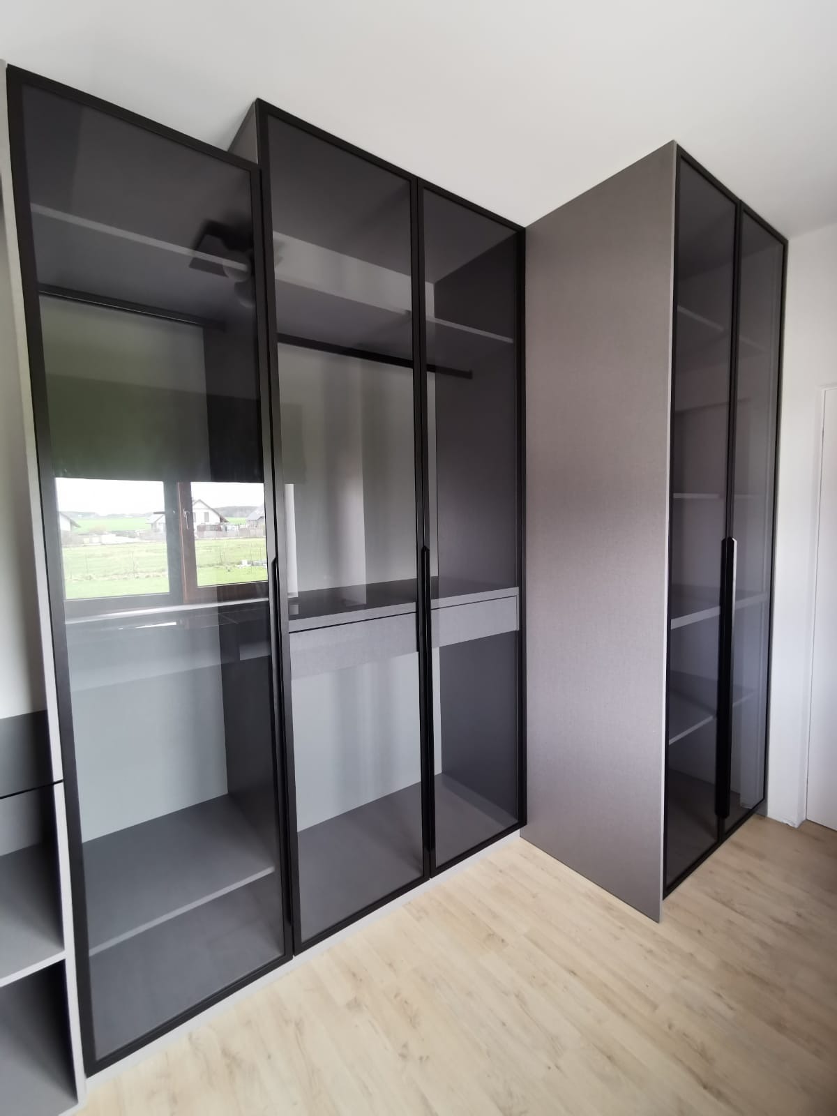 Nowoczesna szafa wnękowa z przesuwnymi, szklanymi drzwiami w czarnej ramie, widziana w jasnym wnętrzu w Kobierzycach. Z okna odbija się widok na wiejski krajobraz.