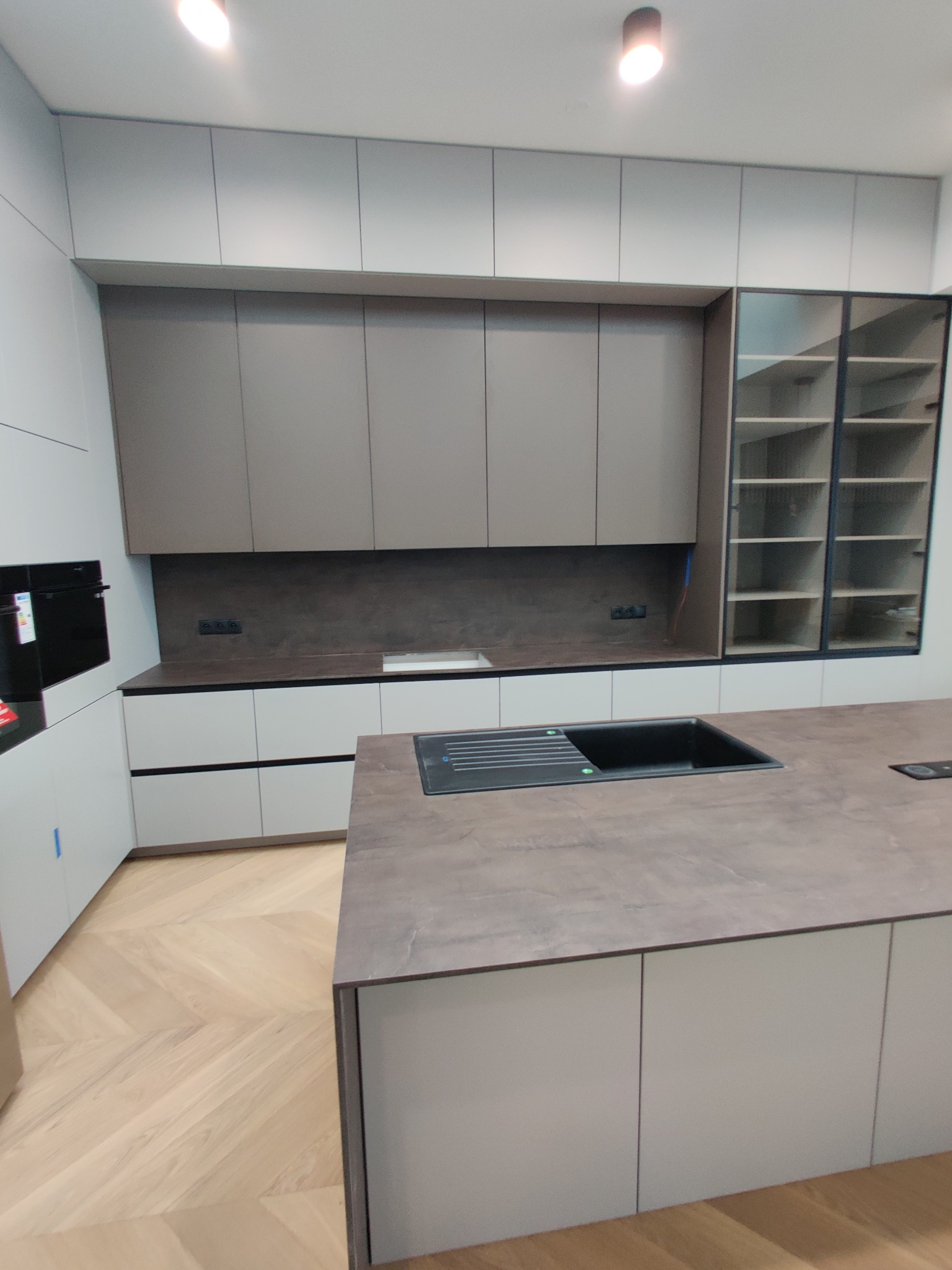 Nowoczesna kuchnia z wyspą, fronty w odcieniach szarości i bieli, parkiet w jodełkę, czarny zlew wkomponowany w blat wyspy, przeszklona szafka z półkami.