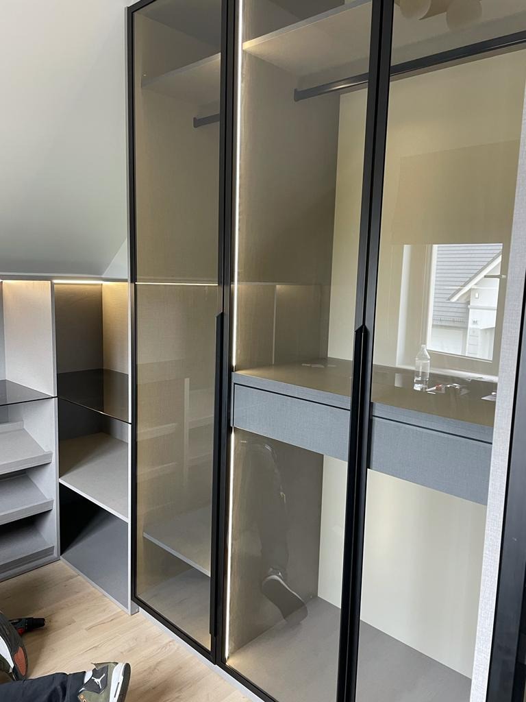 Nowoczesna szafa wnękowa z przesuwnymi drzwiami ze szkła dymionego i oświetleniem LED, widoczne półki i szuflady wewnątrz, fragment podłogi z butami pracownika w dolnym rogu.