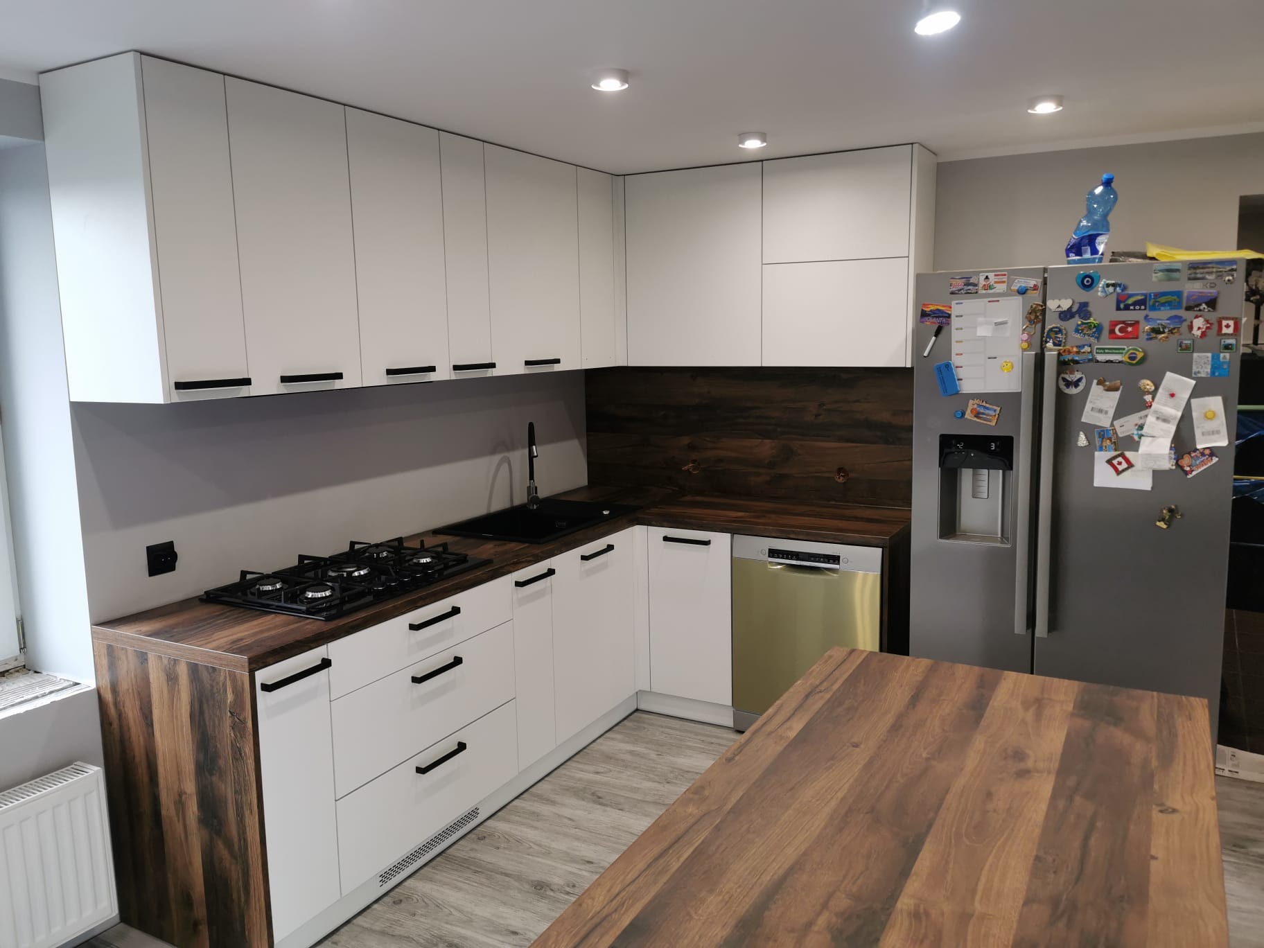 Nowoczesna kuchnia z białymi frontami szafek i blatem imitującym ciemne drewno, czarne uchwyty, płyta gazowa i srebrna lodówka z magnesami.