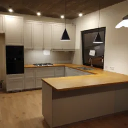 Jasnoszara zabudowa kuchenna z drewnianym blatem, czarnym piekarnikiem i płytą gazową, oświetlona wiszącymi lampami w kształcie stożka.