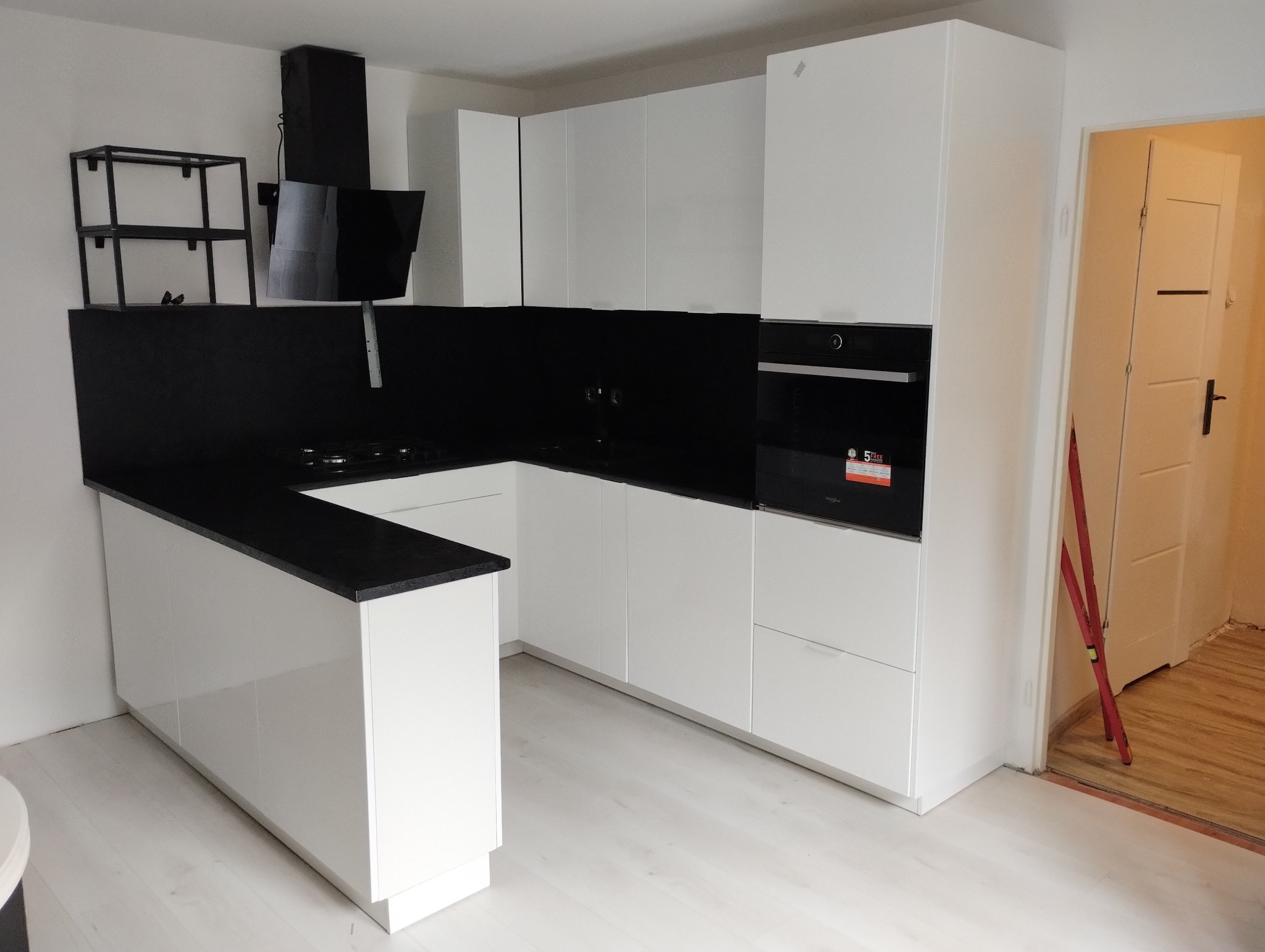 Nowoczesna kuchnia z białymi szafkami i czarnym blatem oraz panelem nad blatem, widoczny czarny okap i półka w tle, drzwi do innego pomieszczenia z boku.