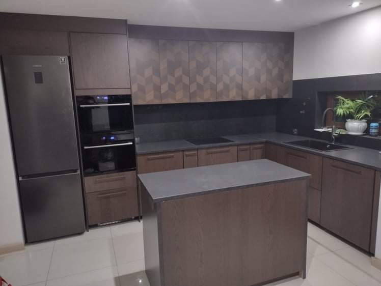 Nowoczesna kuchnia z wyspą, fronty w odcieniach brązu z geometrycznym wzorem na górnych szafkach, sprzęt AGD w zabudowie, widoczny fragment okna z rośliną doniczkową.