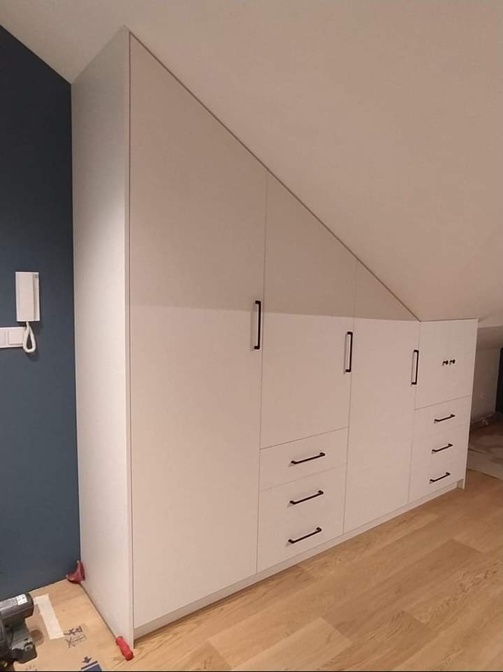 Biała zabudowa meblowa pod skosem, z czarnymi uchwytami, dopasowana do skośnego sufitu, widoczne narzędzia na podłodze, telefon na ścianie.