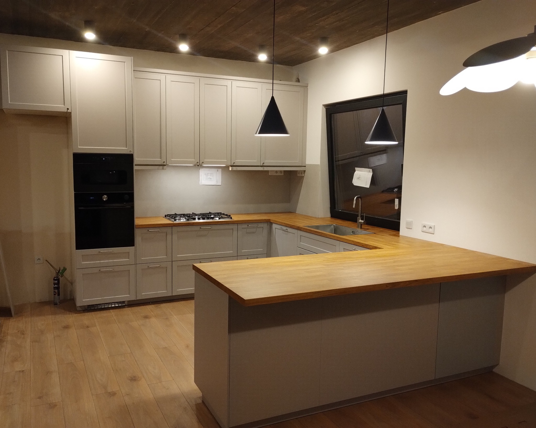 Jasnoszara zabudowa kuchenna z drewnianym blatem, czarnym piekarnikiem i płytą gazową, oświetlona wiszącymi lampami w kształcie stożka.